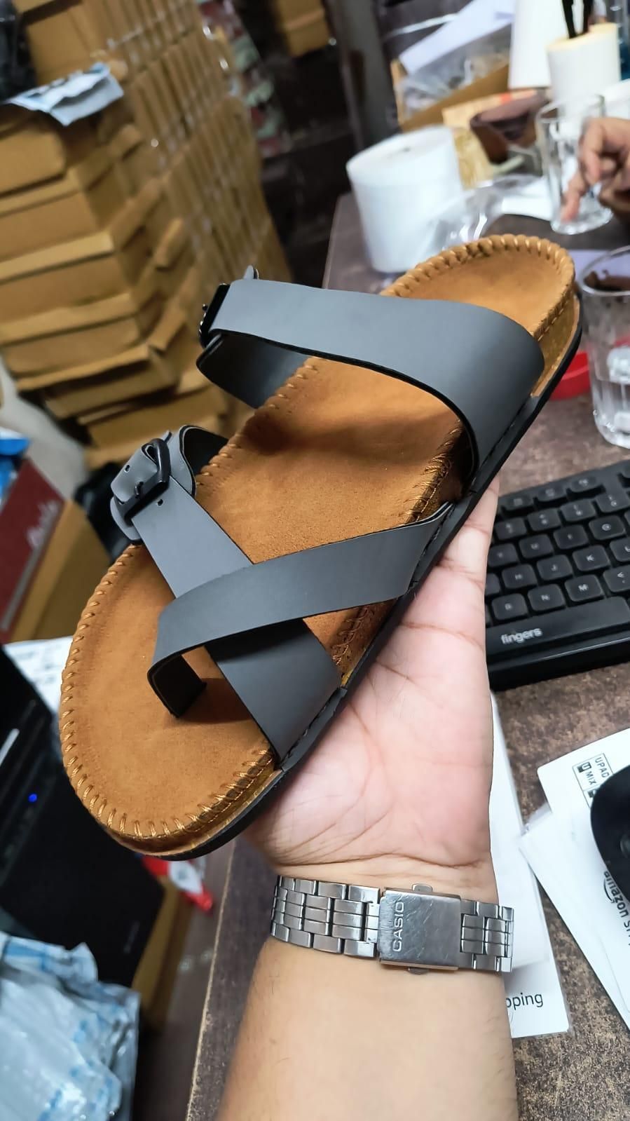 Person holding a sandal with a brown sole and black straps in an indoor setting.
