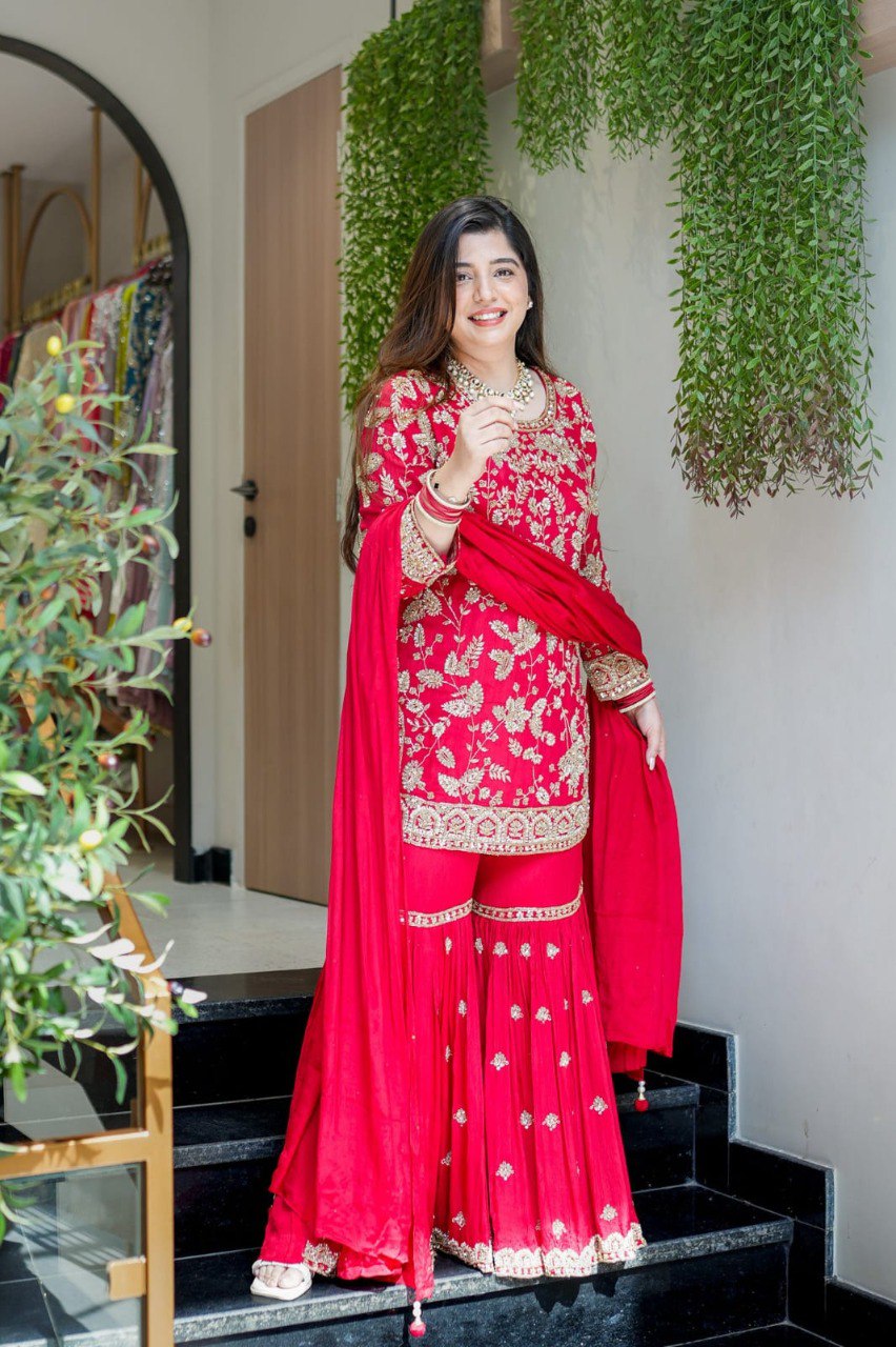 Model smiling in a red embroidered faux georgette sharara suit, standing on stairs, showcasing the full outfit.