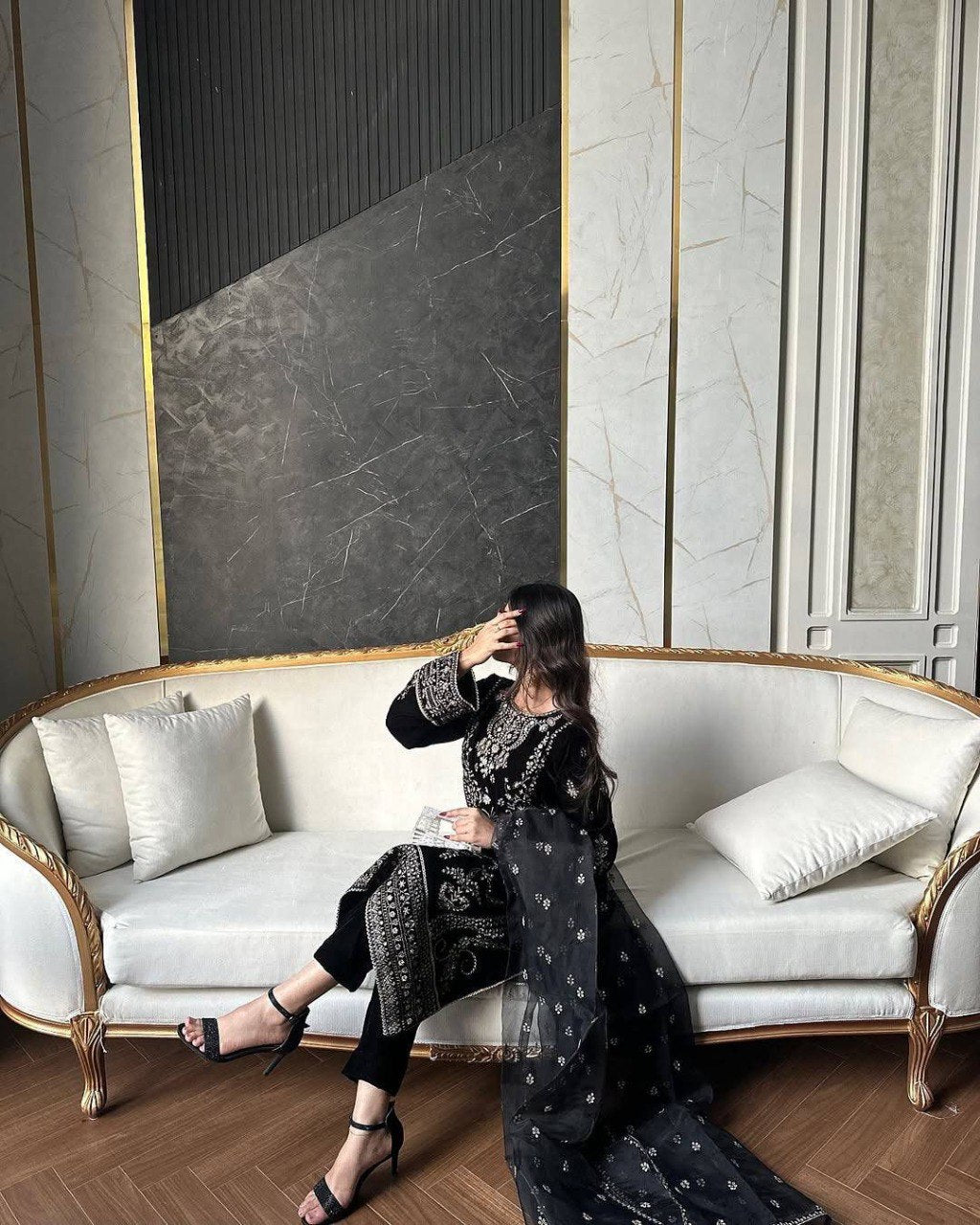 Model in black velvet kurti pant set with intricate embroidery, posing on a white sofa.
