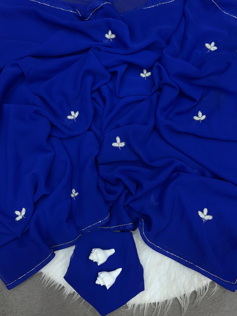 Flat lay of a royal blue soft georgette saree featuring detailed silver cut dana leaf work and border, with matching blouse fabric.