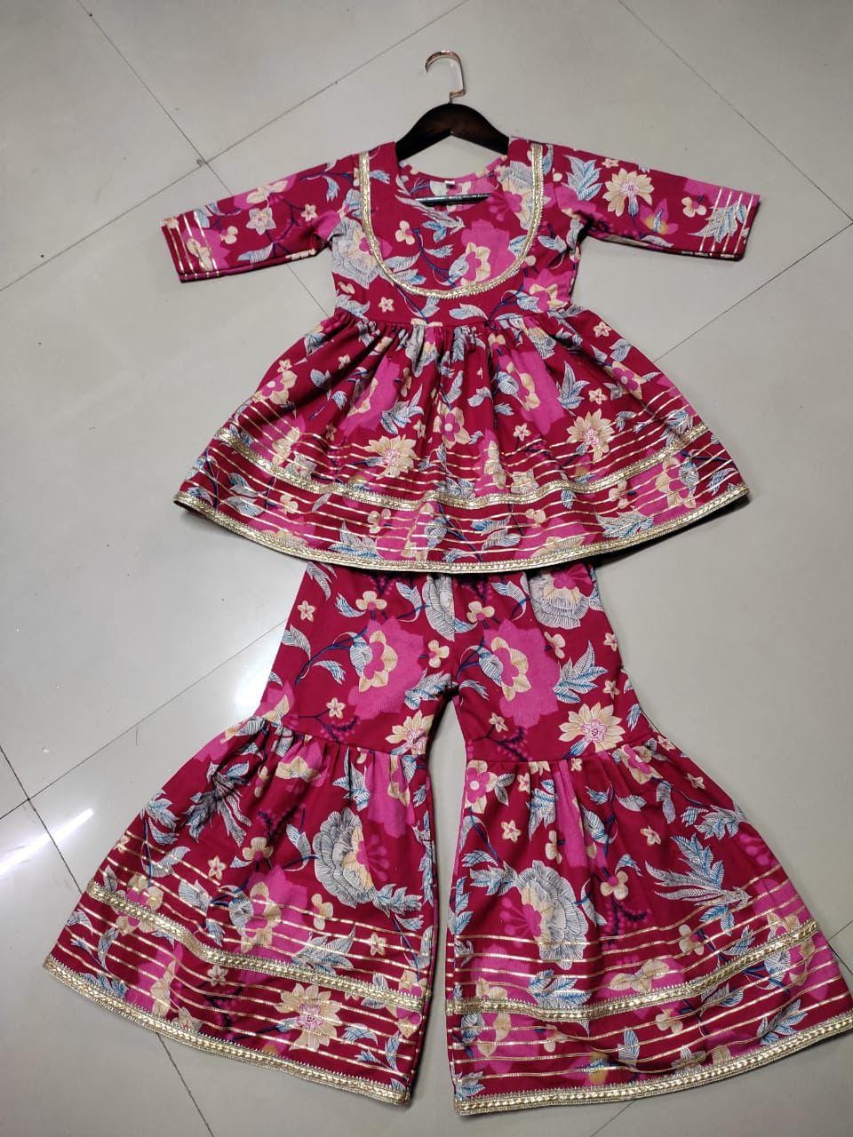 Flat lay of a vibrant maroon floral print Kurta Kediya and Sharara set with gold lace detailing, ready for packaging.