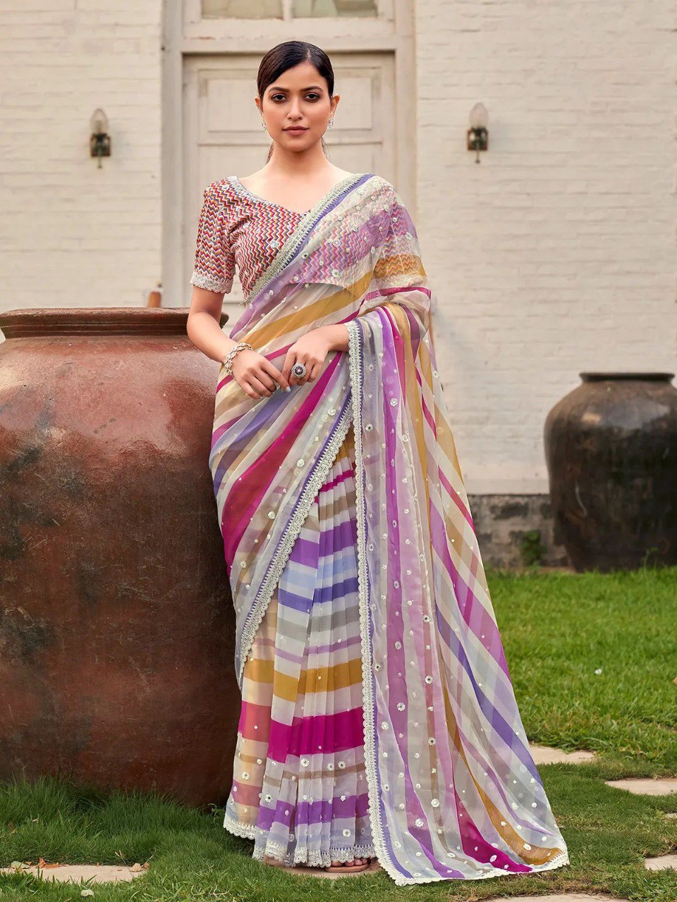 Model standing outdoors in pink and yellow striped saree, displaying full length and drape against a rustic backdrop.