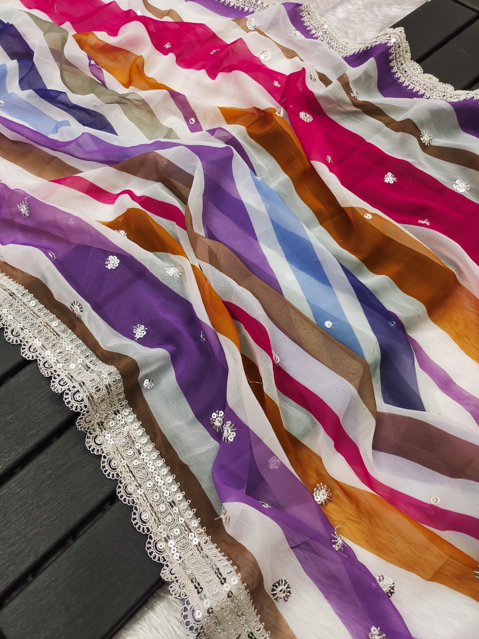 Close-up flat lay of the pink and yellow multi-striped georgette saree fabric with scattered sequins and elaborate cutwork border.