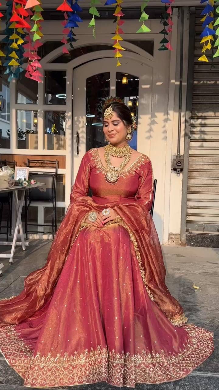 Woman elegantly posing in rust red organza lehenga choli set with heavy gold embroidery and traditional jewelry.