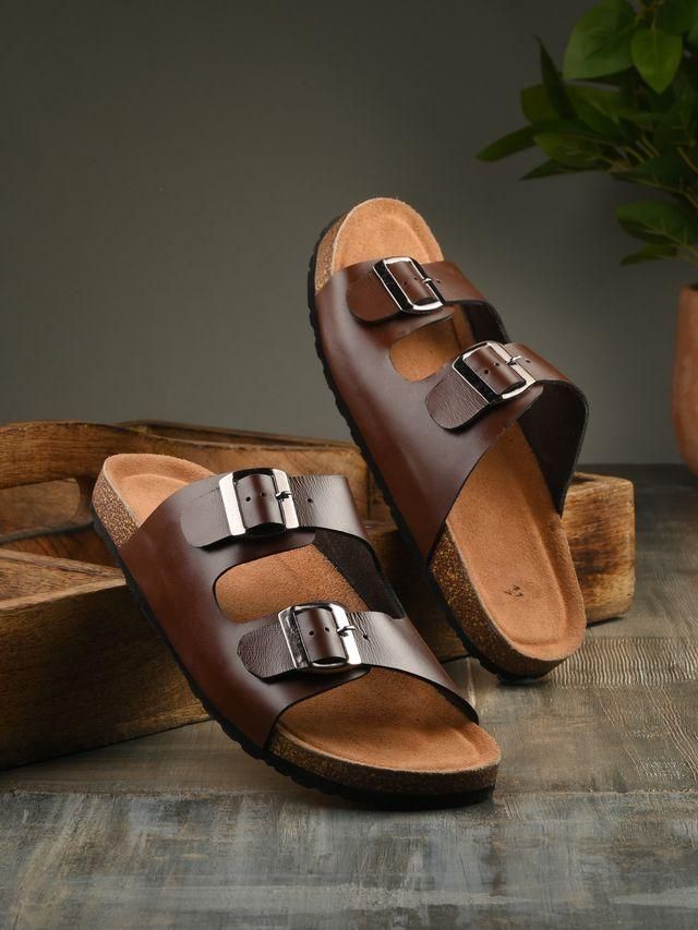 Brown sandals with buckles on a wooden surface with a plant in the background