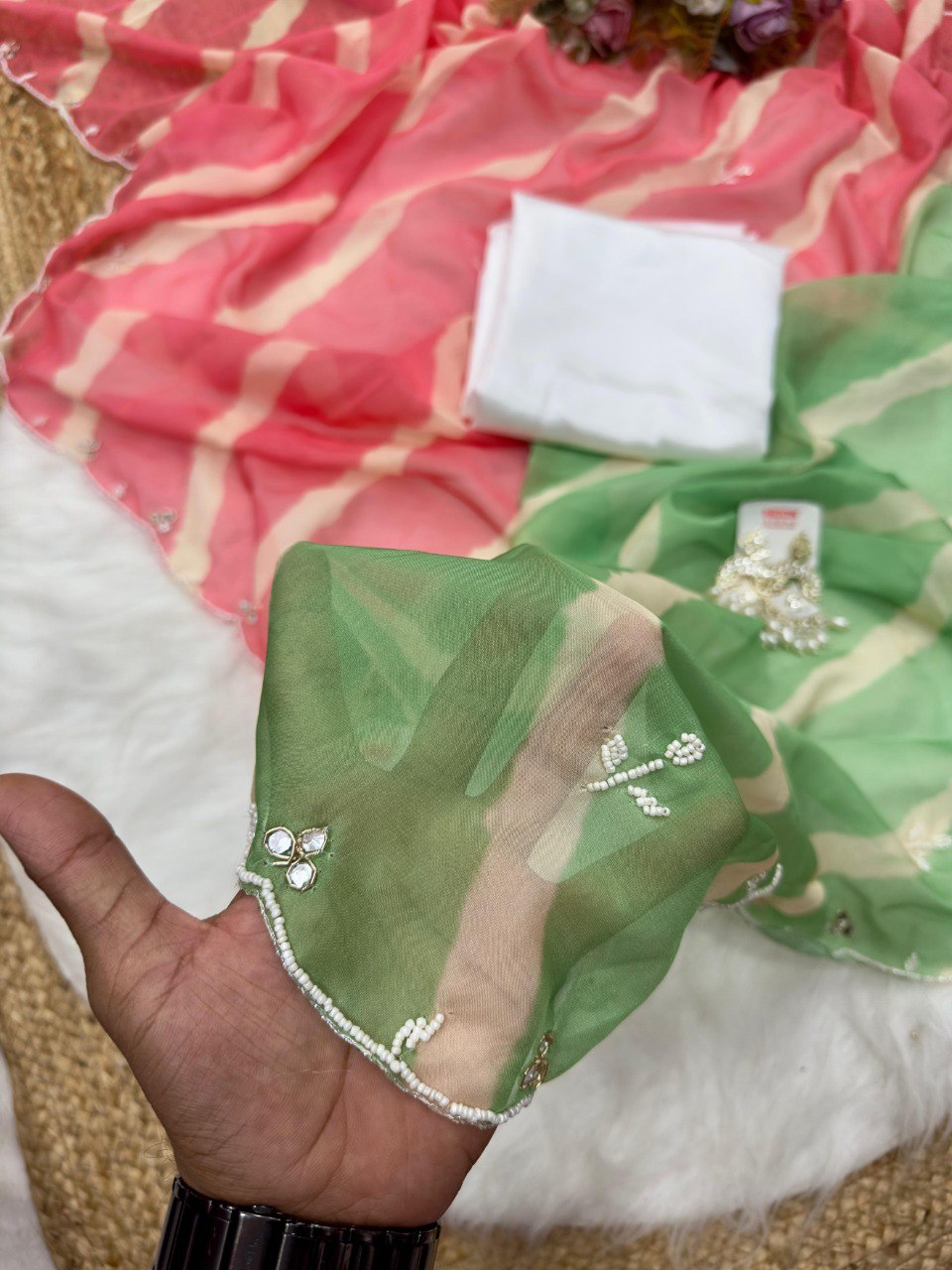 Close-up of green Lehriya Tebi silk fabric swatch with val moti and mirror work border, with pink saree and white blouse piece in background.