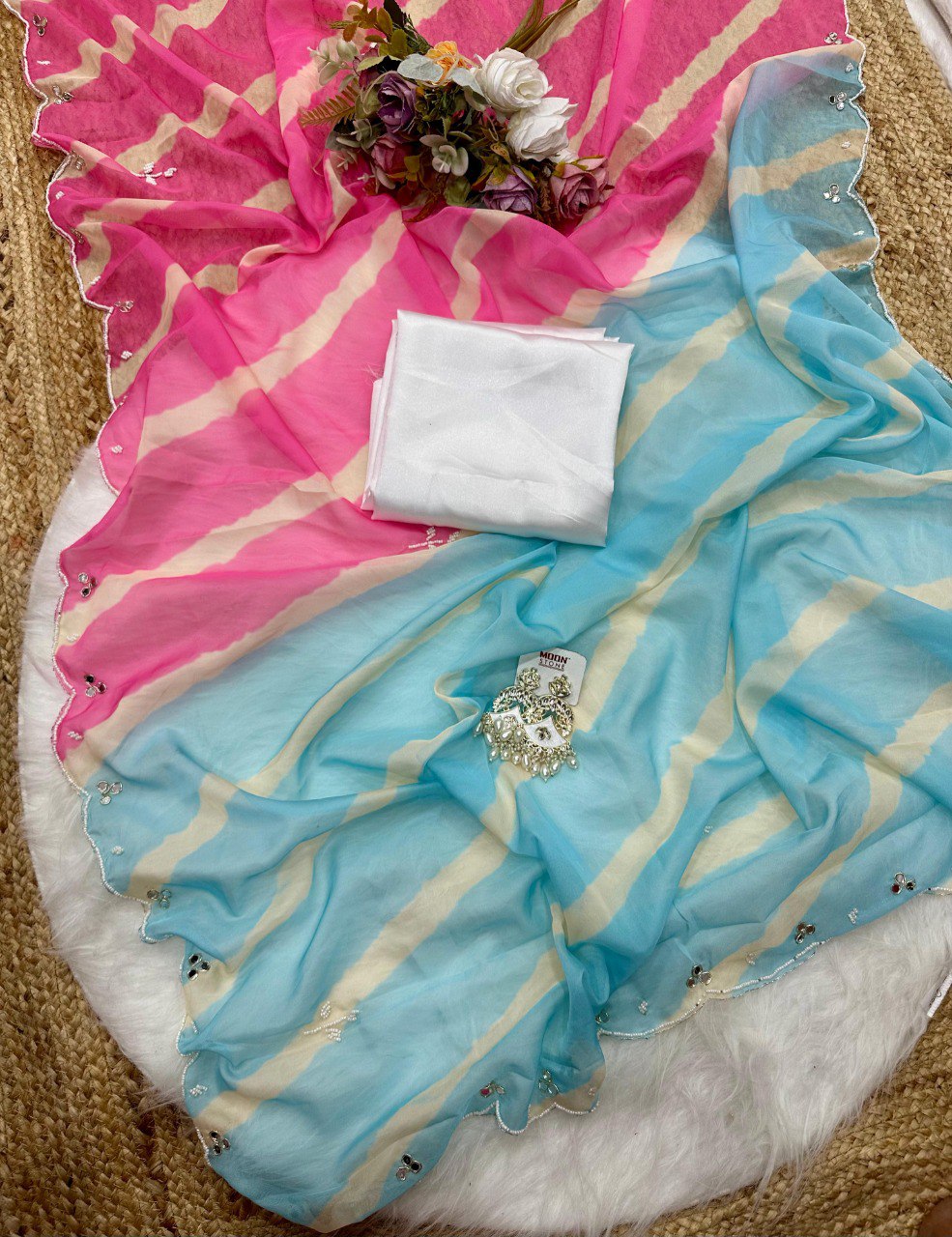 Overhead flat lay of unfolded pink and blue Lehriya Tebi silk saree with handwork, showcasing white blouse piece and earrings.
