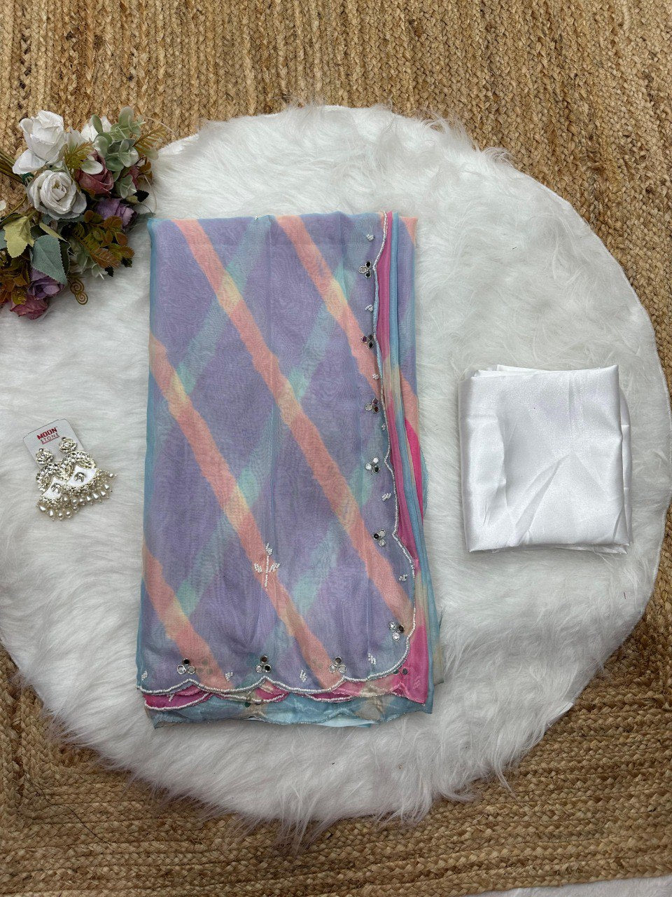 Folded pink and purple Lehriya Tebi silk saree with white blouse piece and designer earrings on a plush white rug.