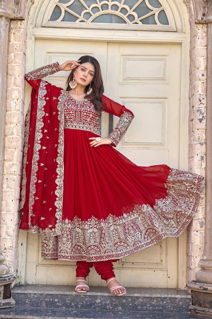 Model posing elegantly in a crimson red heavy faux georgette Anarkali gown with golden sequence embroidery, standing by a vintage door.