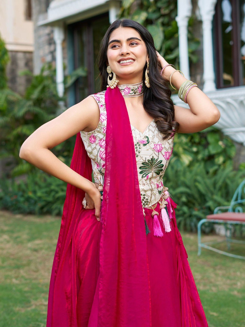 Smiling model wearing a beautiful Rani (magenta) embroidered Lehenga Choli by TRENDBUY, with tassels and a flowing dupatta, posing in a garden.