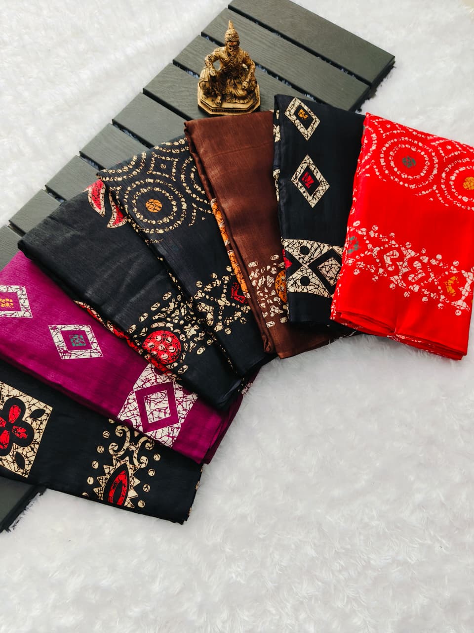 A flat lay display of seven rolled dola silk sarees in various colors and batik patterns, with a decorative idol.