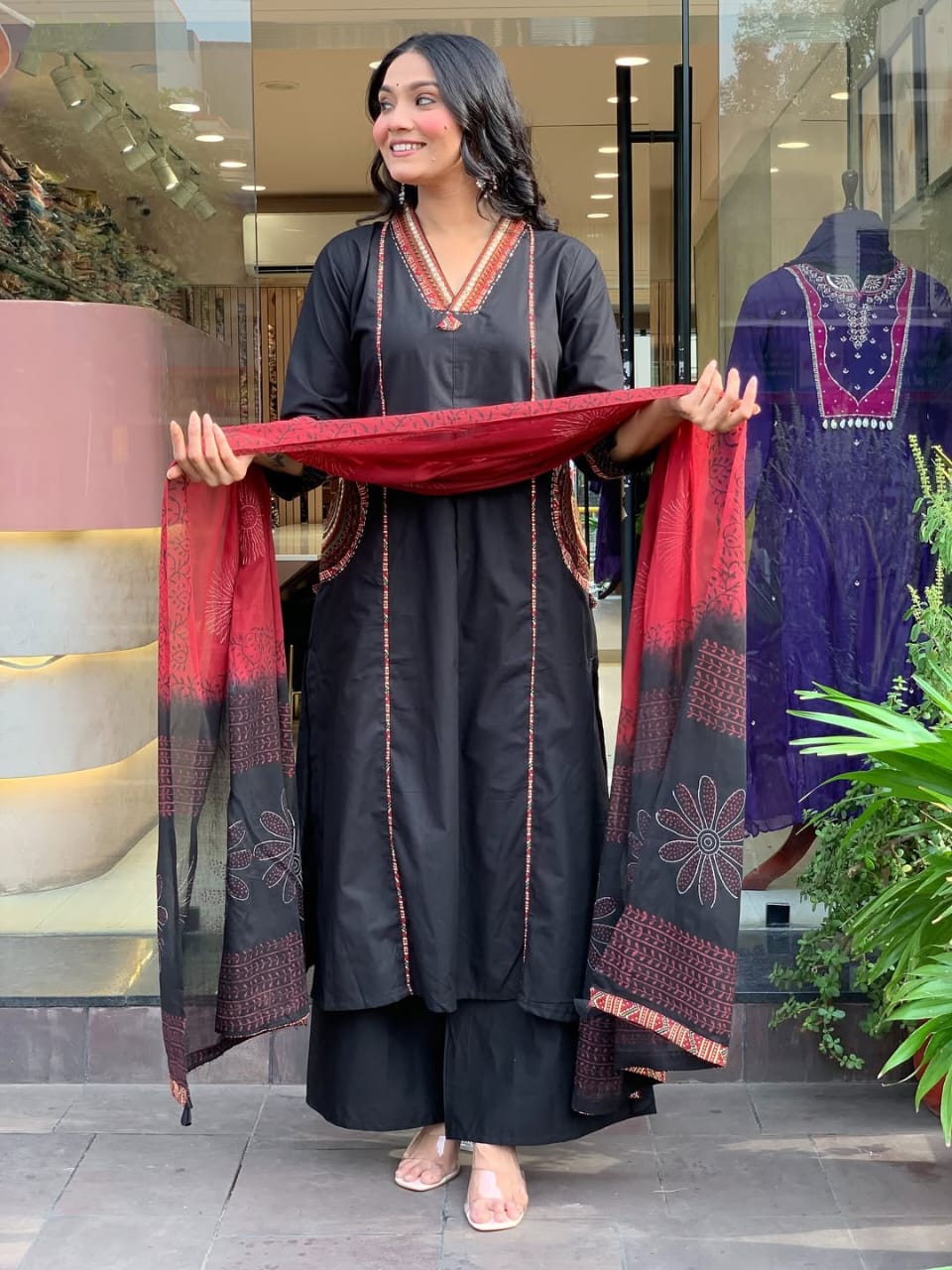 Full shot of a smiling woman in a black kurta set with a red and black patterned dupatta, holding it out.