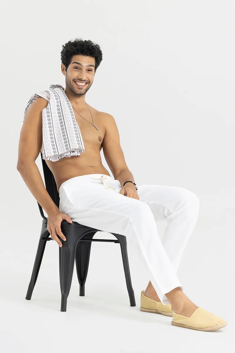 Man sitting on a black chair, smiling, wearing white cotton linen pants and a patterned cloth draped over his shoulder.