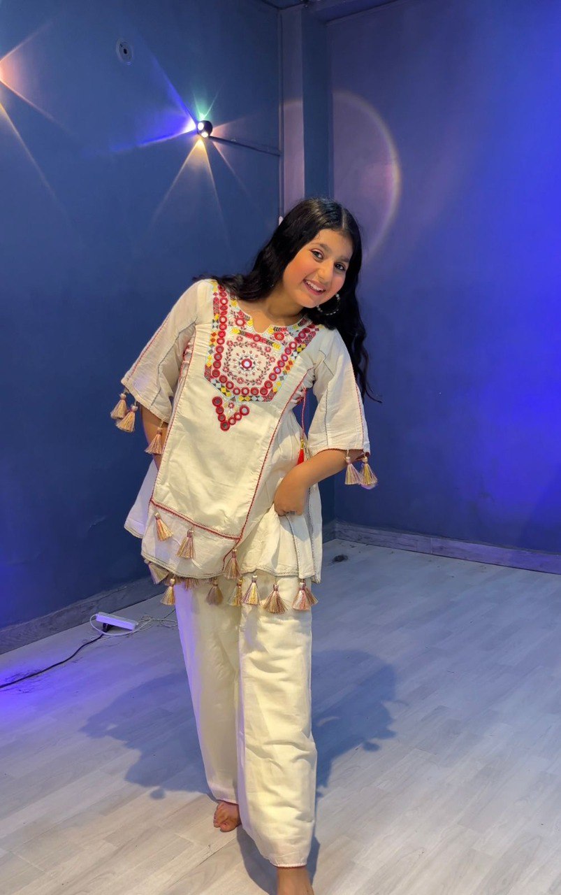 Happy girl in white khadi cotton outfit with colourful embroidery and mirrors, standing casually.