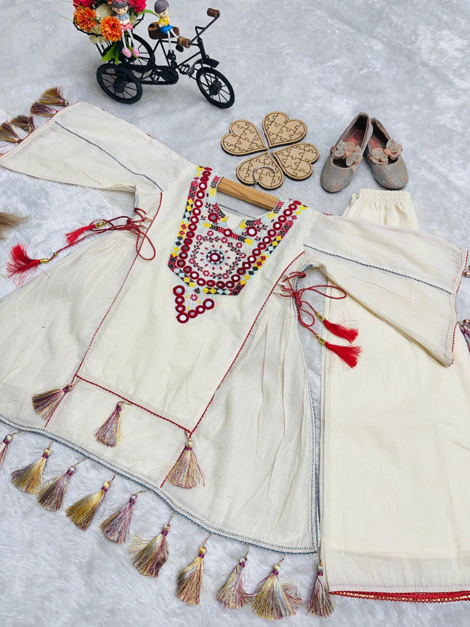 Flat lay of white khadi cotton plazzo set with embroidery, tassels, along with small shoes and decorative items.