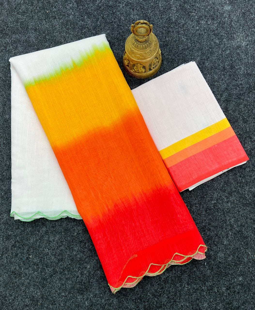 Overhead flat lay displaying the white and multi-color ombre linen saree with its matching blouse fabric, next to a decorative antique bell.