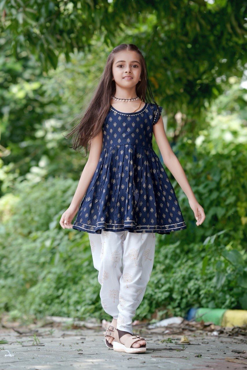 Young girl wearing a navy blue dress with white polka dots and white pants standing outdoors.