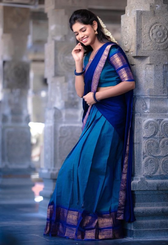 Woman in a blue saree with gold border standing against stone pillars.
