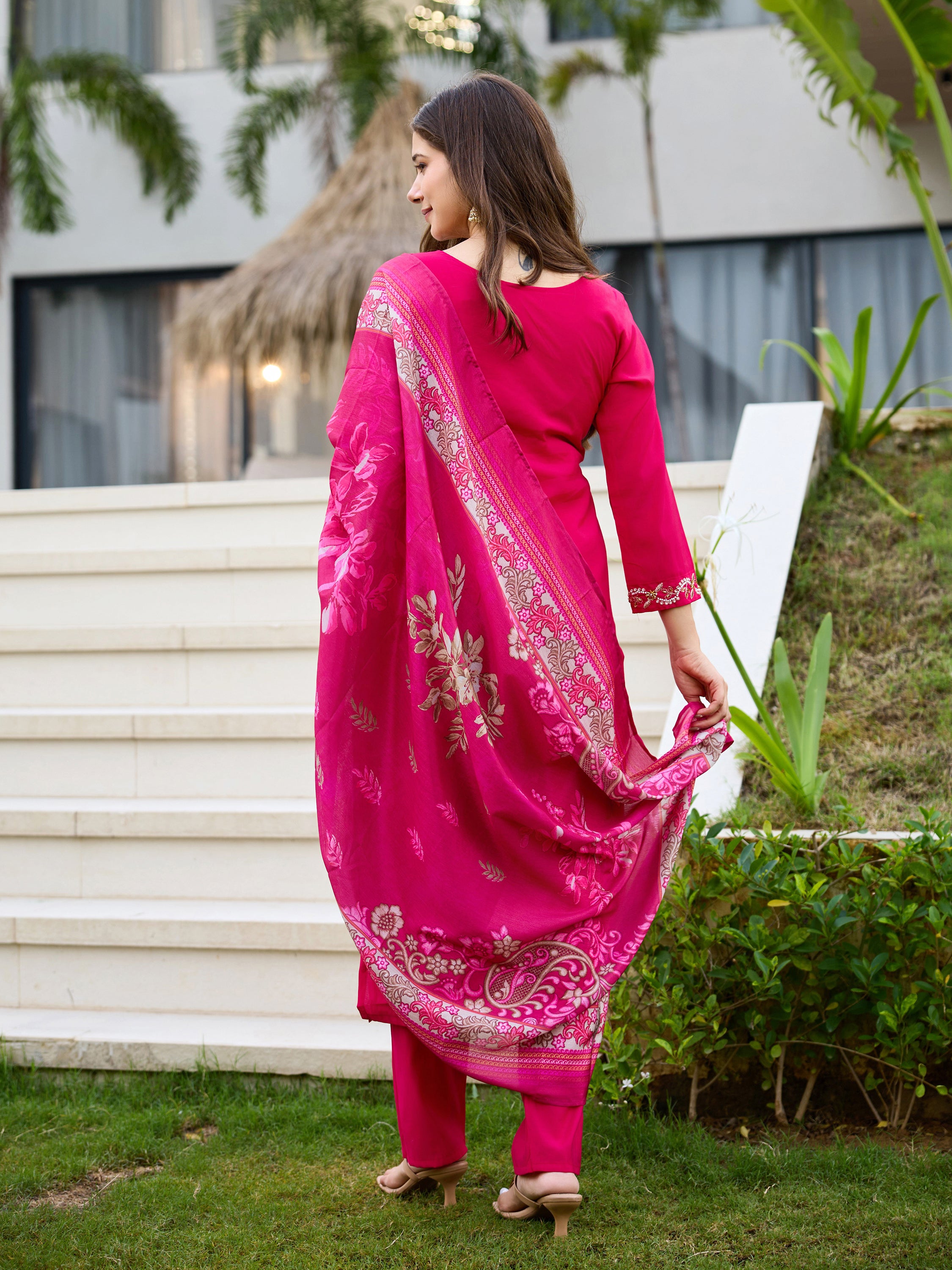 Woman in full pink Roman silk kurti set with floral print dupatta flowing, seen from behind, in an outdoor setting.