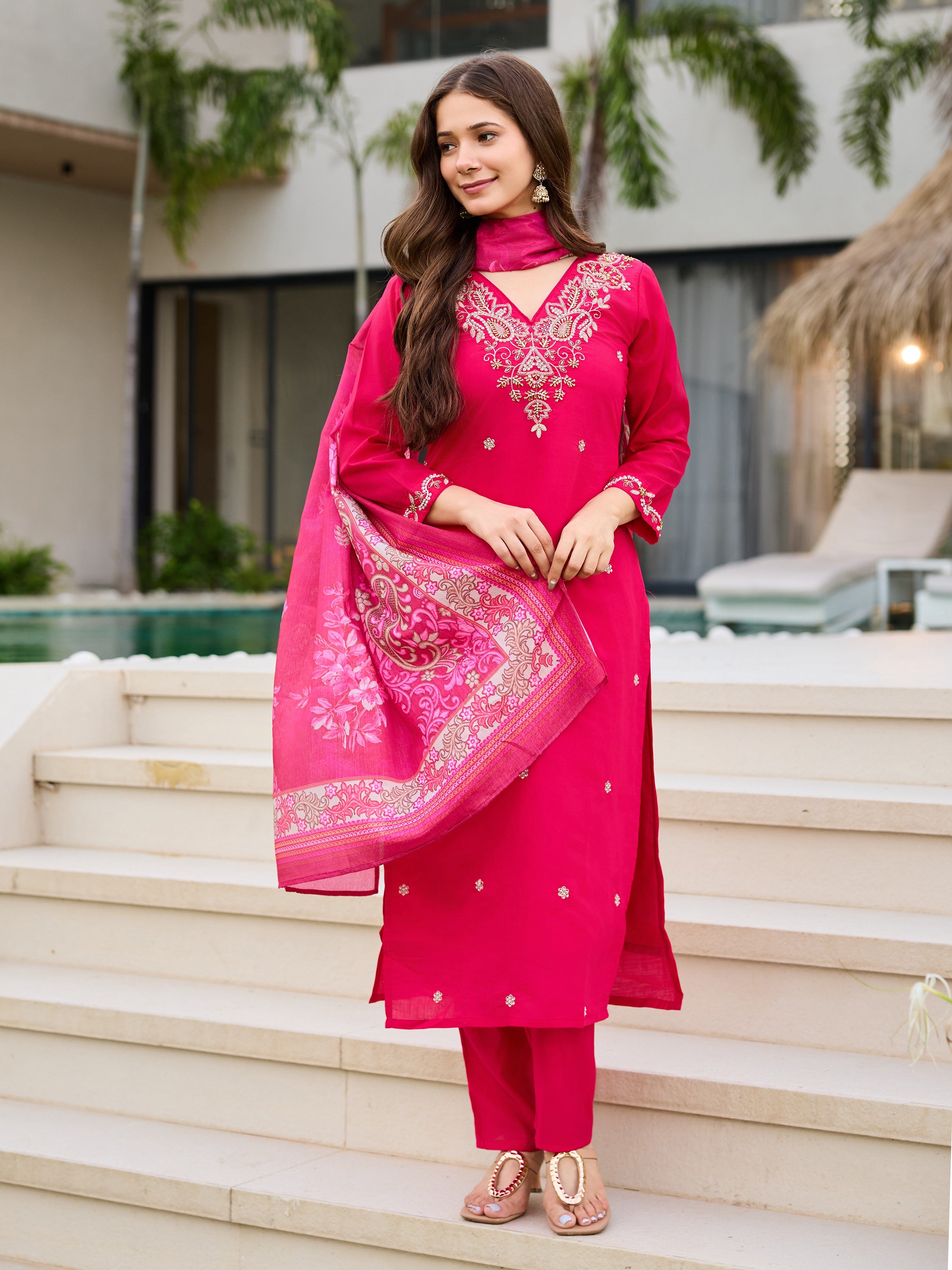 Full body shot of a woman in the complete pink kurti, pant, and dupatta set, holding the dupatta, elegant pose against white stairs.