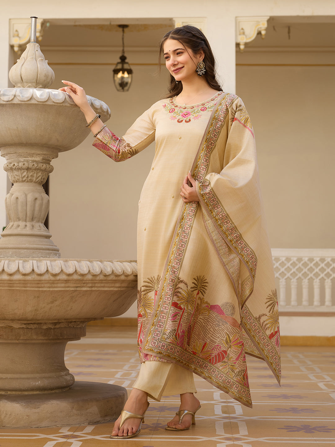 Model posing gracefully in a cream Viscose Assam Silk Kurta Set with embroidered neckline and printed dupatta, beside an ornate fountain.