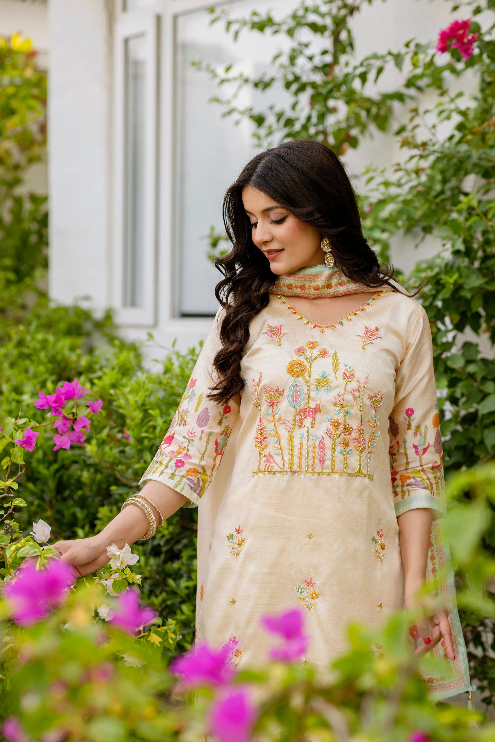 Model admiring flowers, wearing TRENDBUY Viscose Assam Silk Kurta Set, emphasizing the vibrant embroidered patterns and bead details on the kurta.