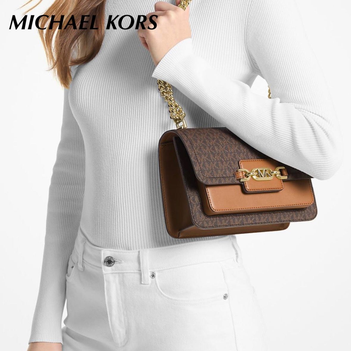 Person holding a brown handbag with gold chain strap, wearing a white sweater and jeans, on a plain background.