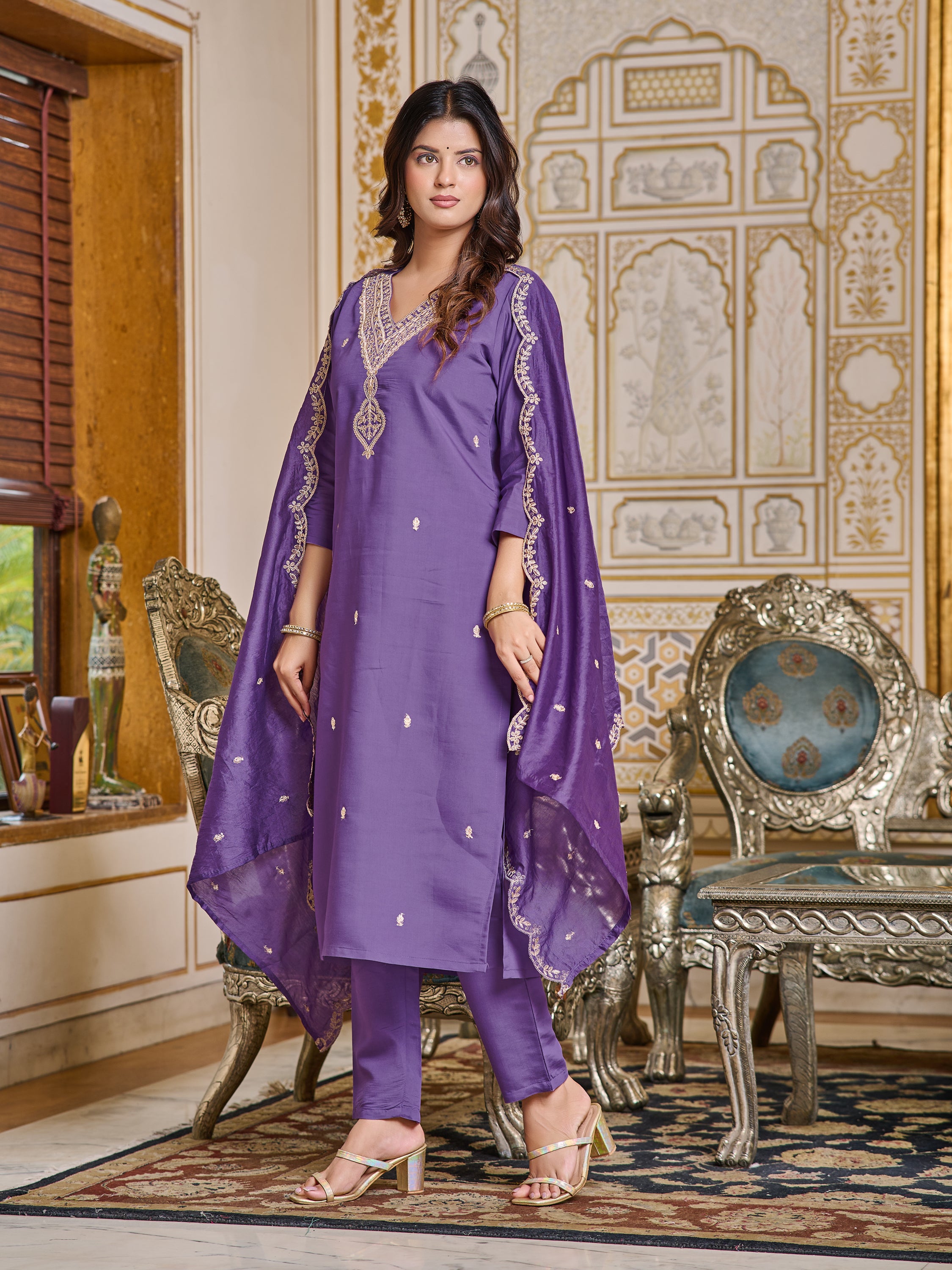 Woman in purple kurti set, with dupatta draped, standing in a regal interior.