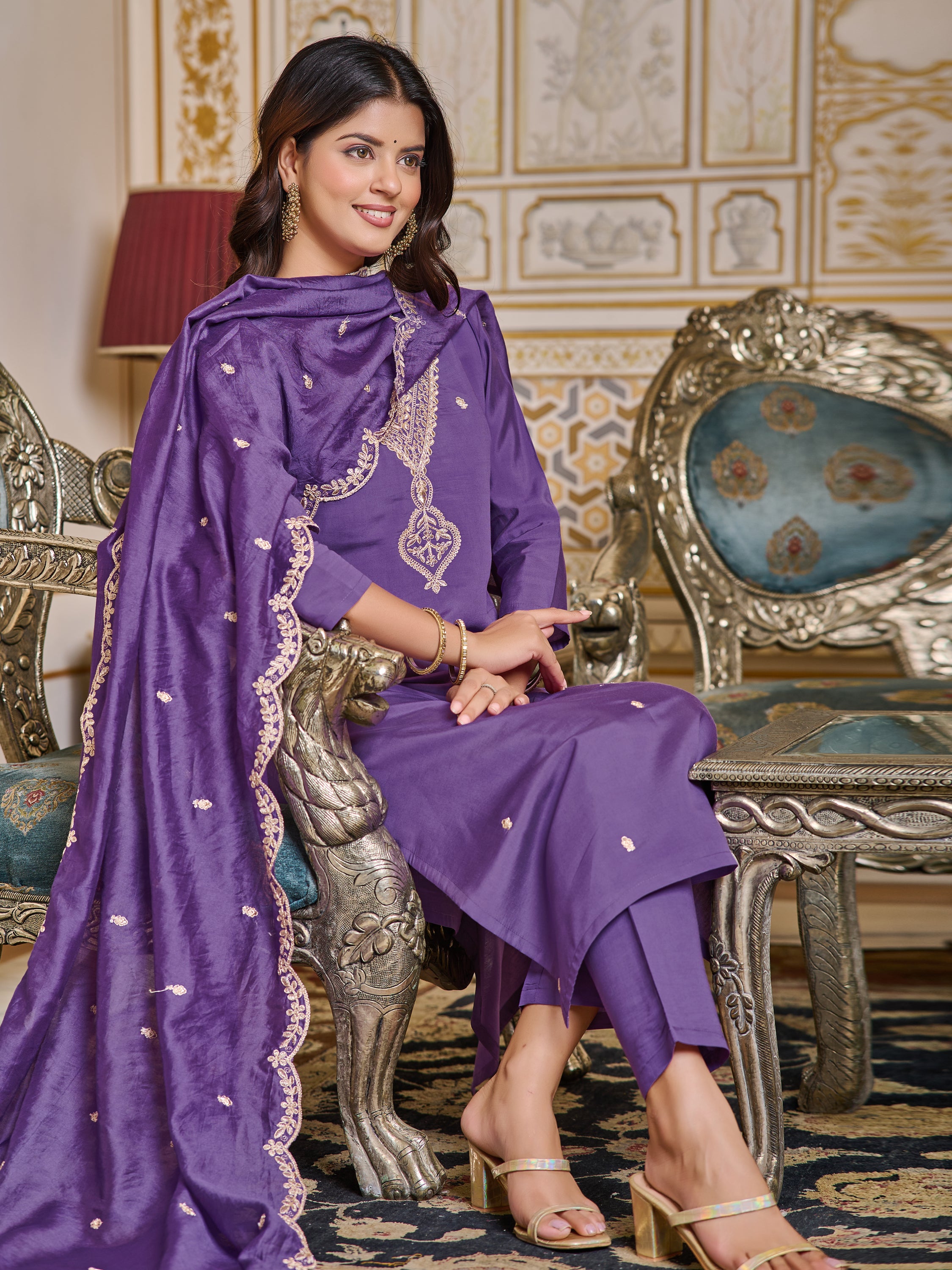 Woman sitting on an ornate chair, smiling, showcasing the comfort and style of the purple chanderi set.