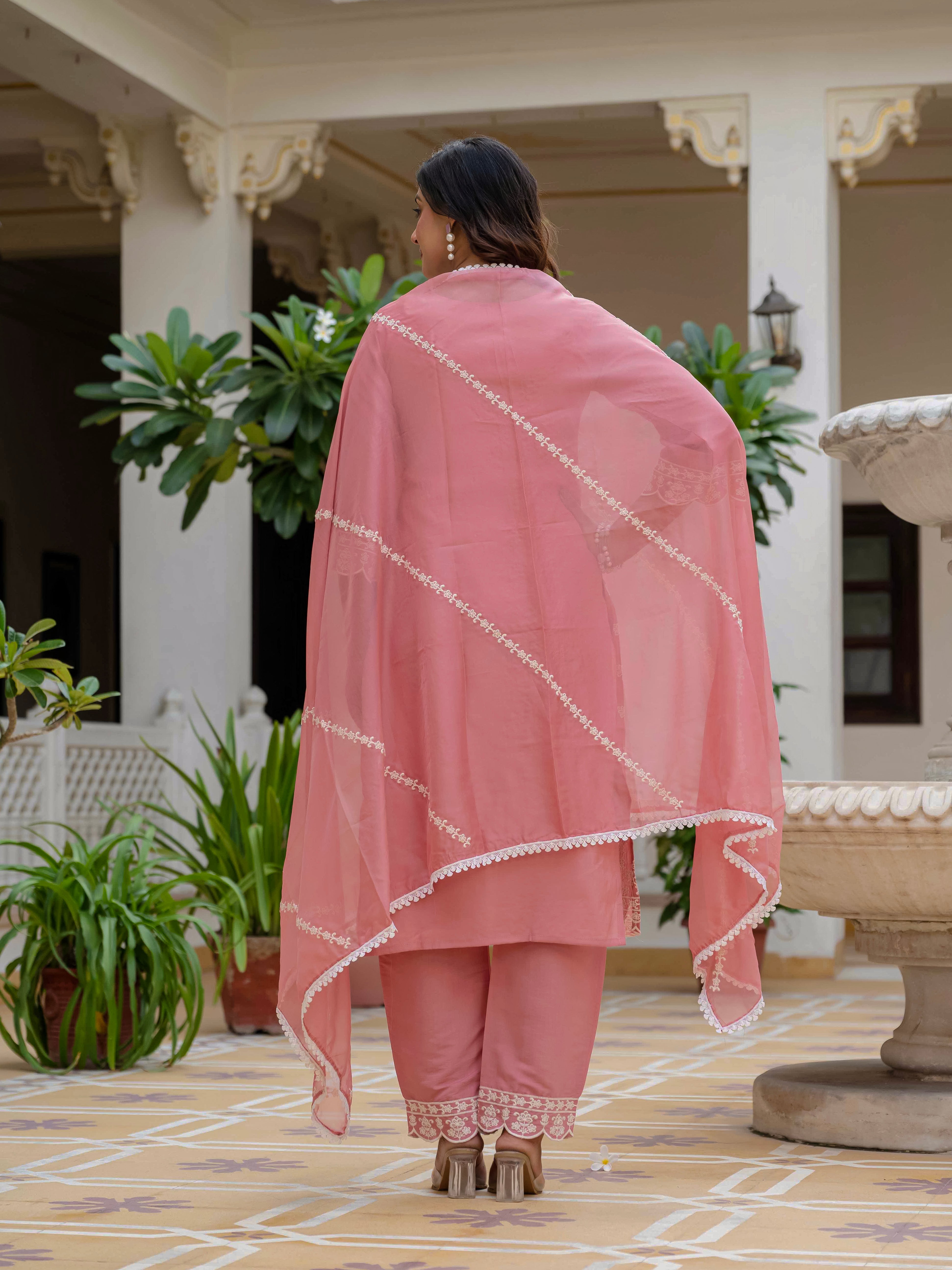 Back view of a woman in a rose pink Roman silk kurti set, highlighting the flowing organza dupatta with white lace trim and embroidered pant hem.