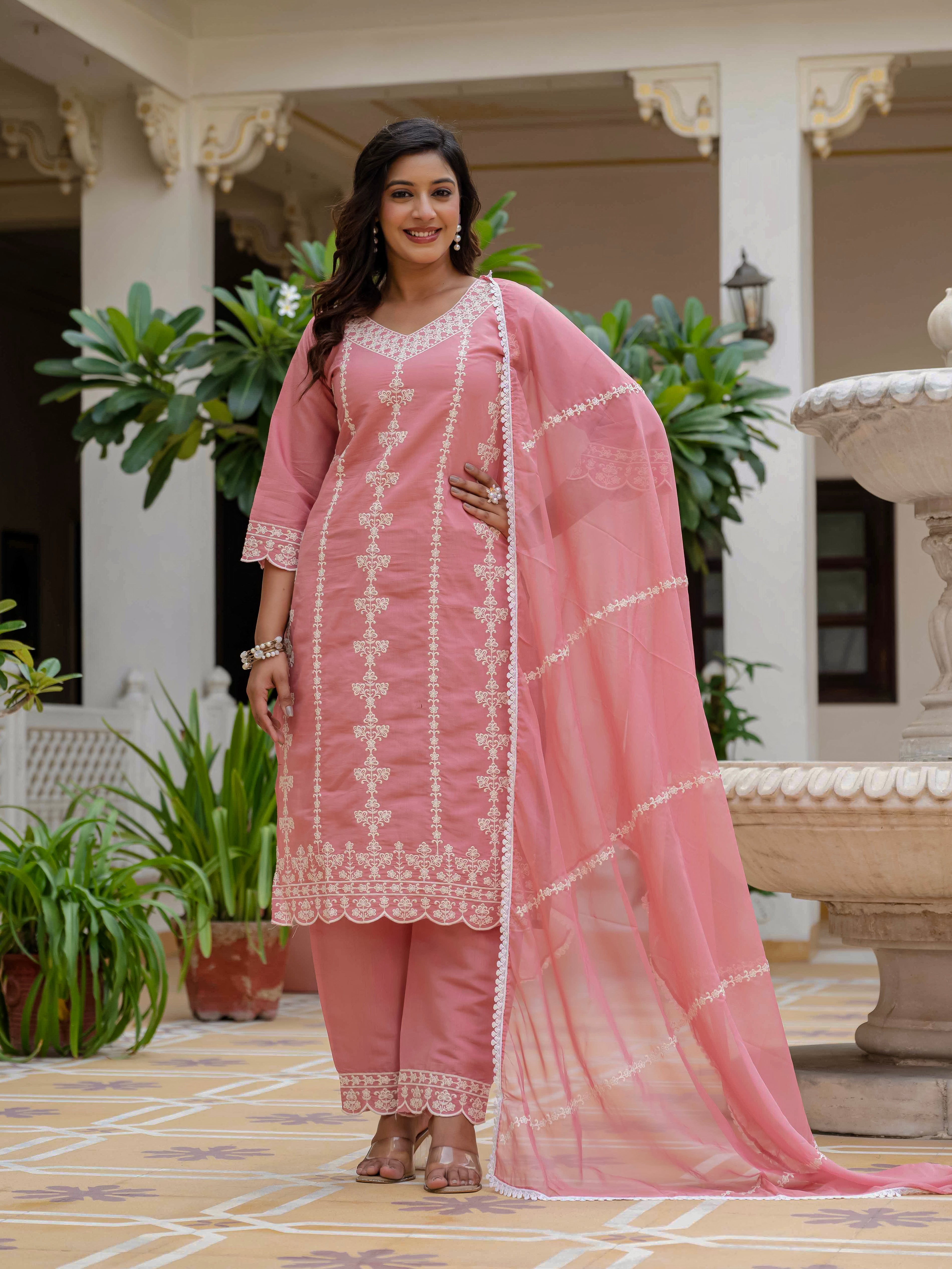 Full shot of a smiling woman in a rose pink Roman silk embroidered kurti, matching pant, and organza dupatta, posing elegantly outdoors.