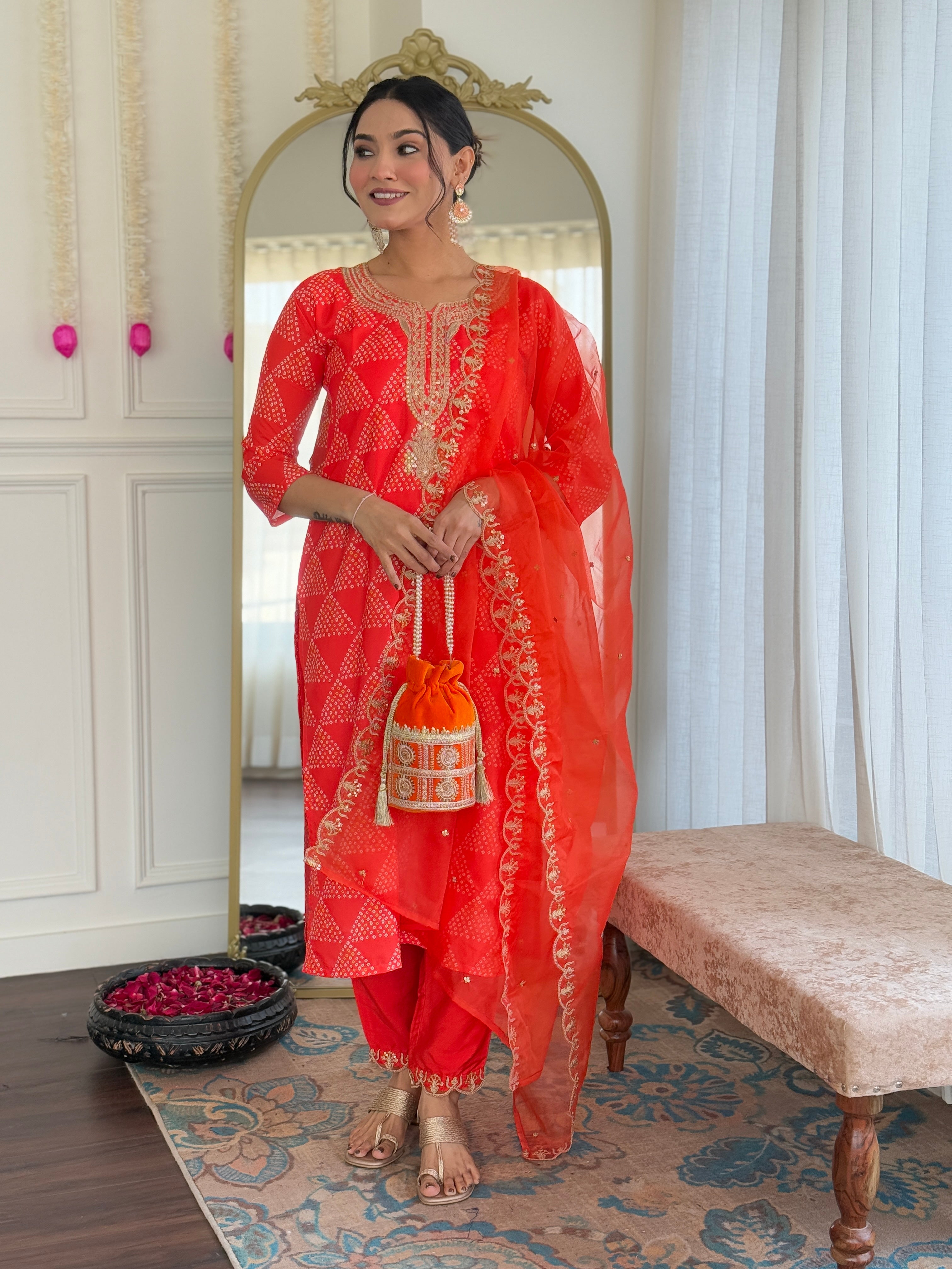 Full-length shot of a woman in a vibrant orange embroidered kurti set with a decorative potli bag, standing in an elegant room.