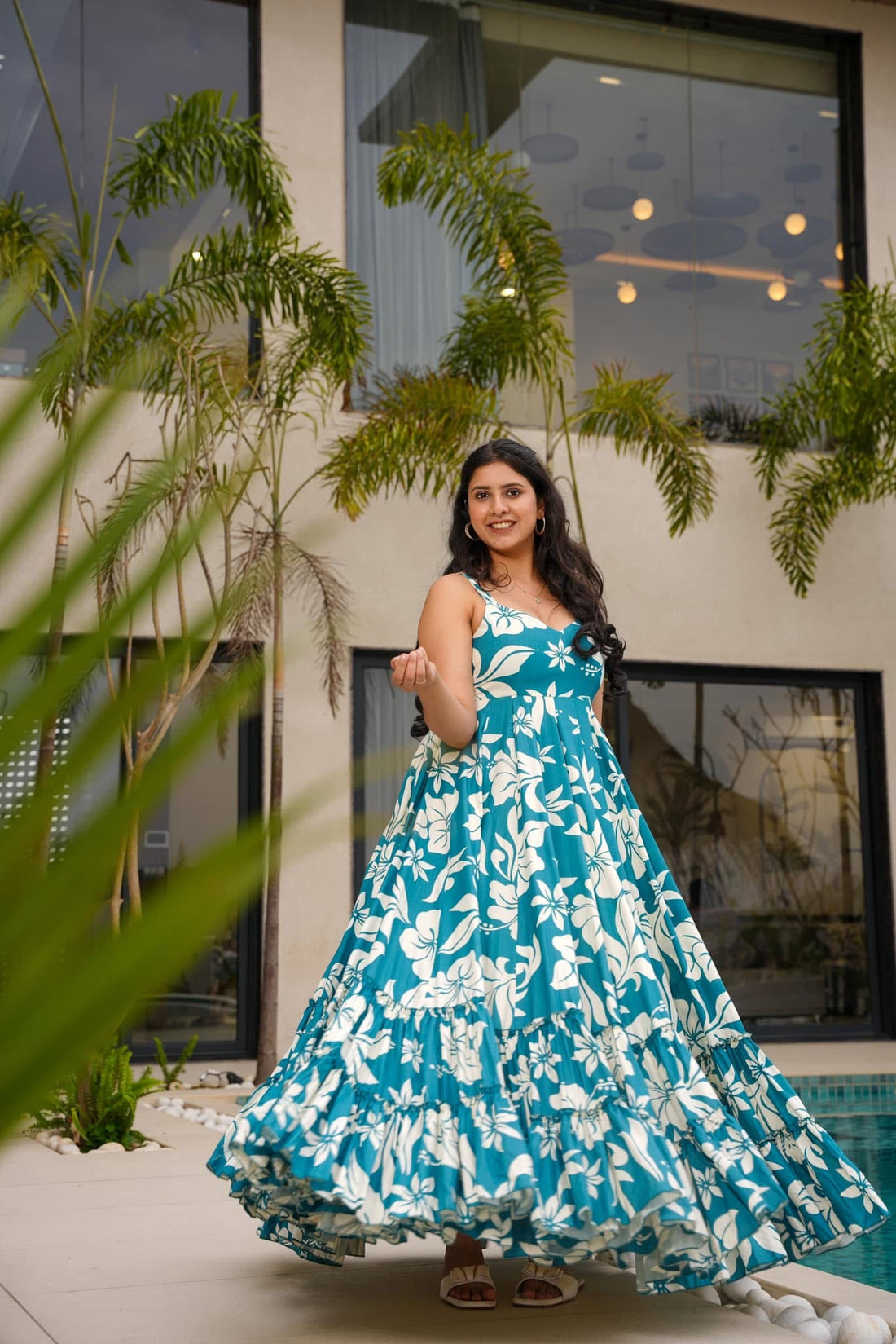 Smiling woman in a flowing teal and white floral tiered maxi dress, posing elegantly by a pool, enjoying a sunny day.