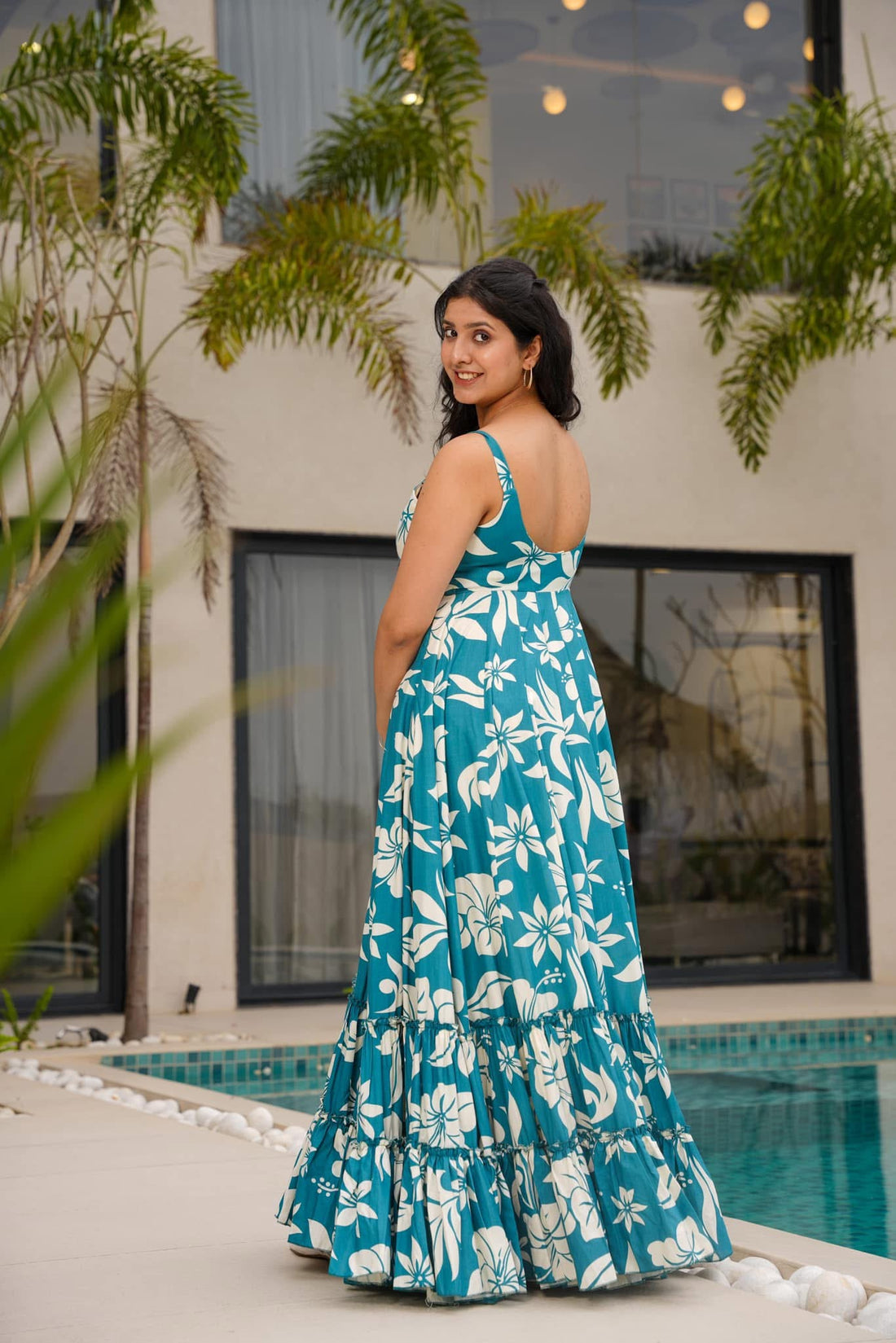 Woman in a stylish teal floral print maxi dress looking back over her shoulder, showing the graceful flow of the tiered skirt outdoors.