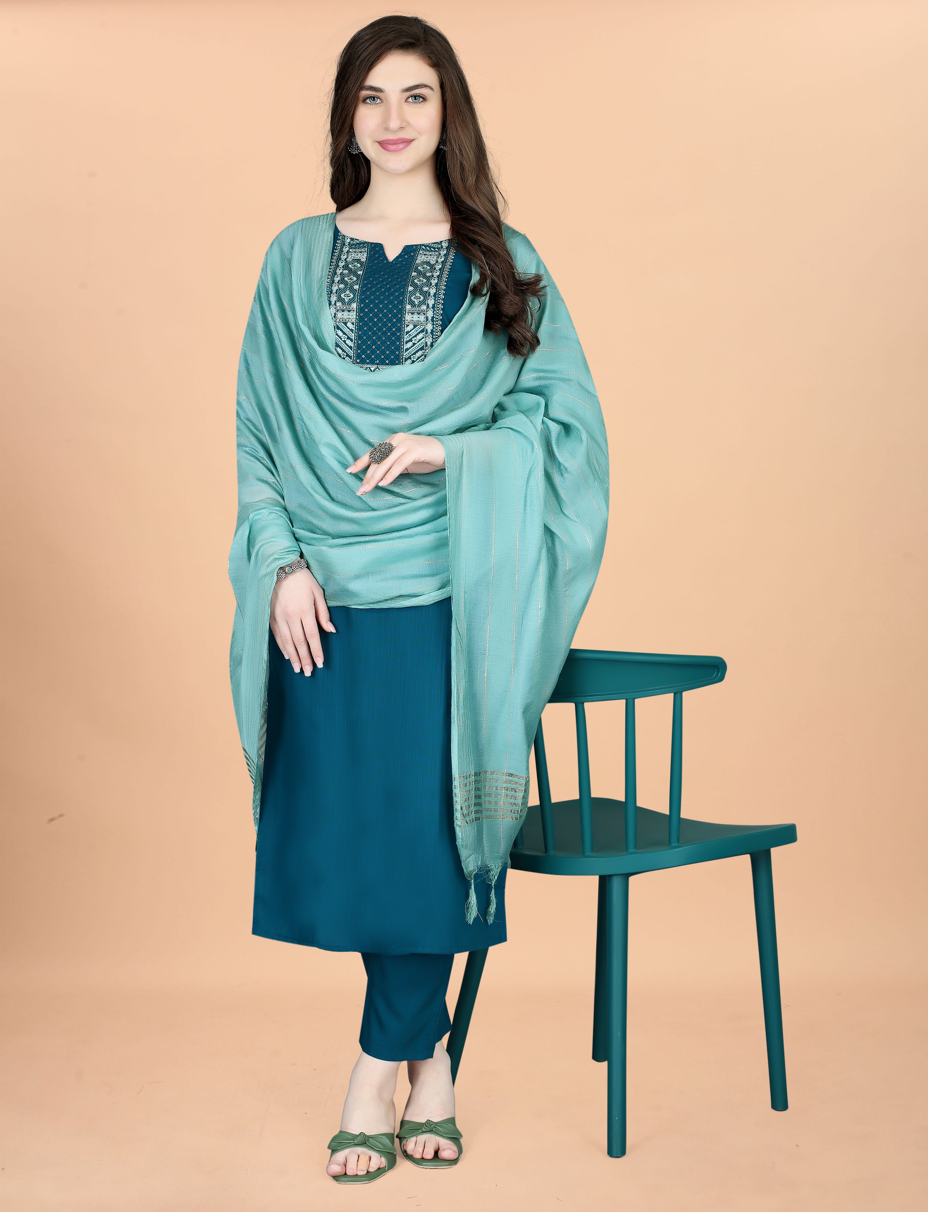 Full front view of a woman posing with a matching teal chair, wearing the embroidered teal kurti set with a gracefully draped mint green dupatta.