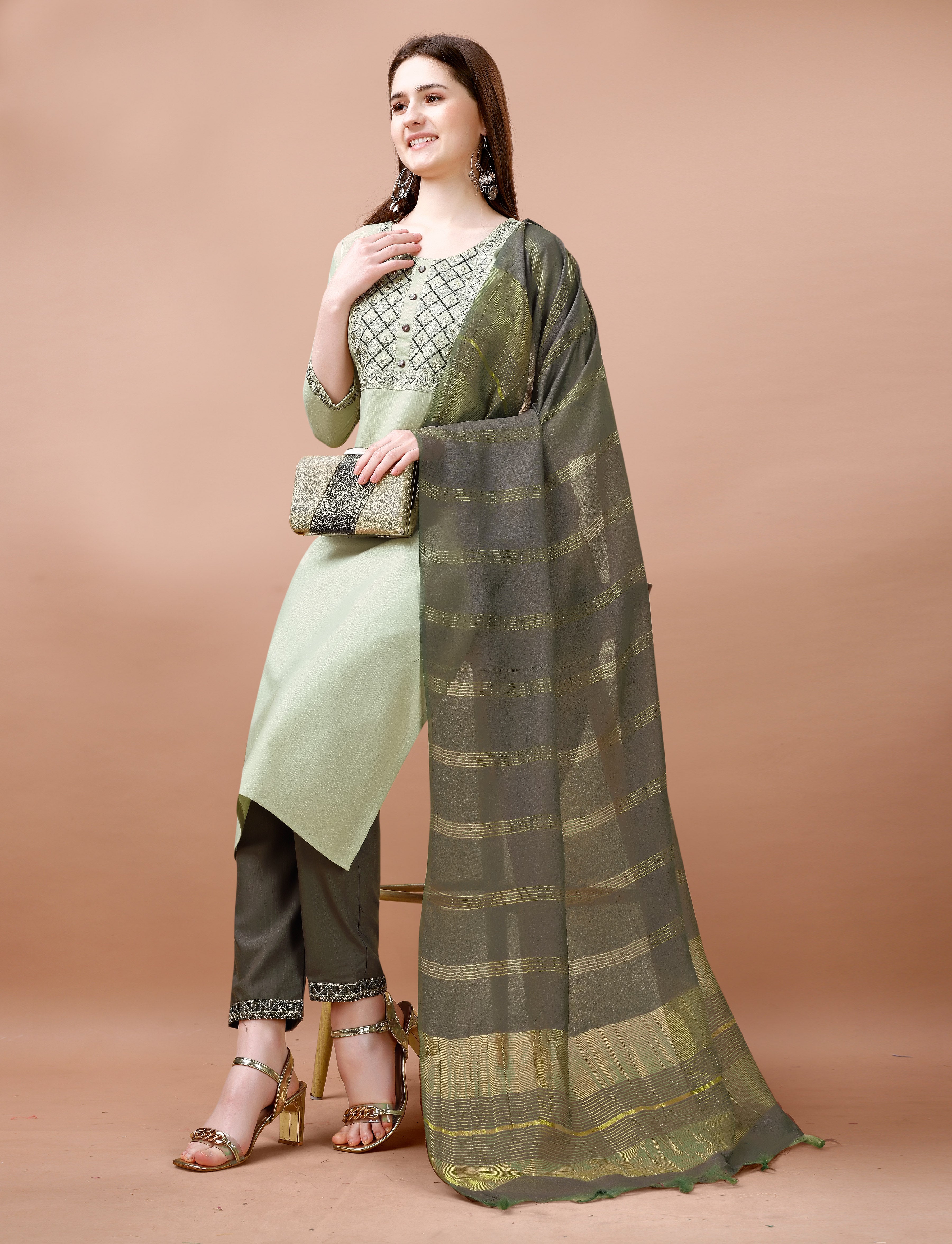Woman in a sage green embroidered kurta set, sitting on a stool, showcasing the ensemble, embroidered pant cuffs, and golden heels.