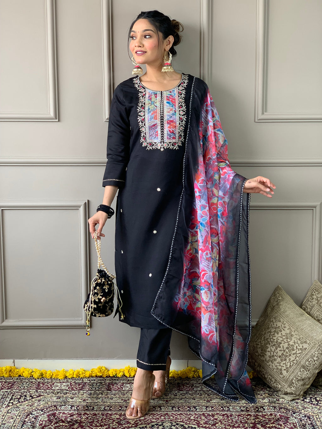 Full shot of a woman in TRENDBUY black embroidered kurta set, holding a black potli bag, with a vibrant floral organza dupatta draped over her shoulder.