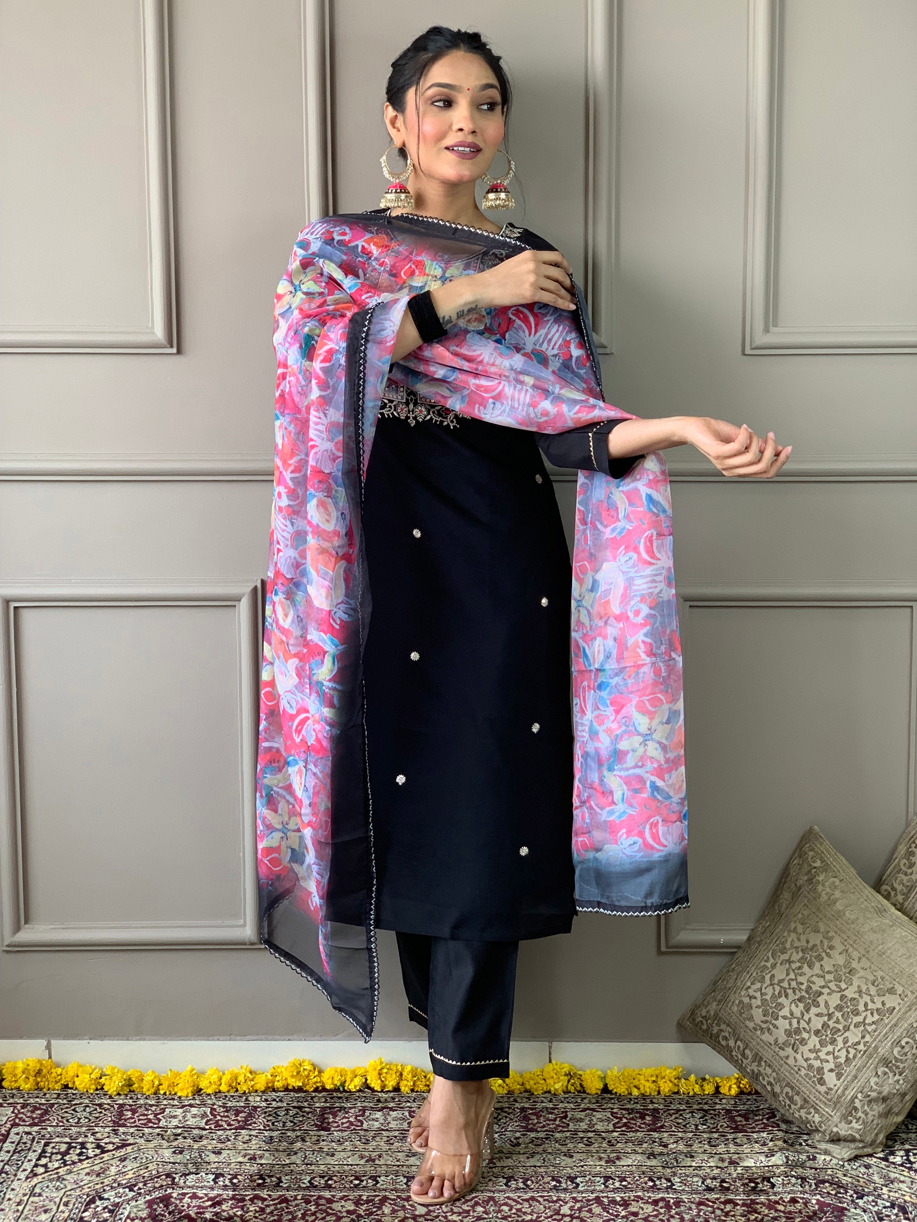 Woman gracefully posing in TRENDBUY black embroidered kurti, matching pants, and flowing colorful organza dupatta against a light grey panelled wall.