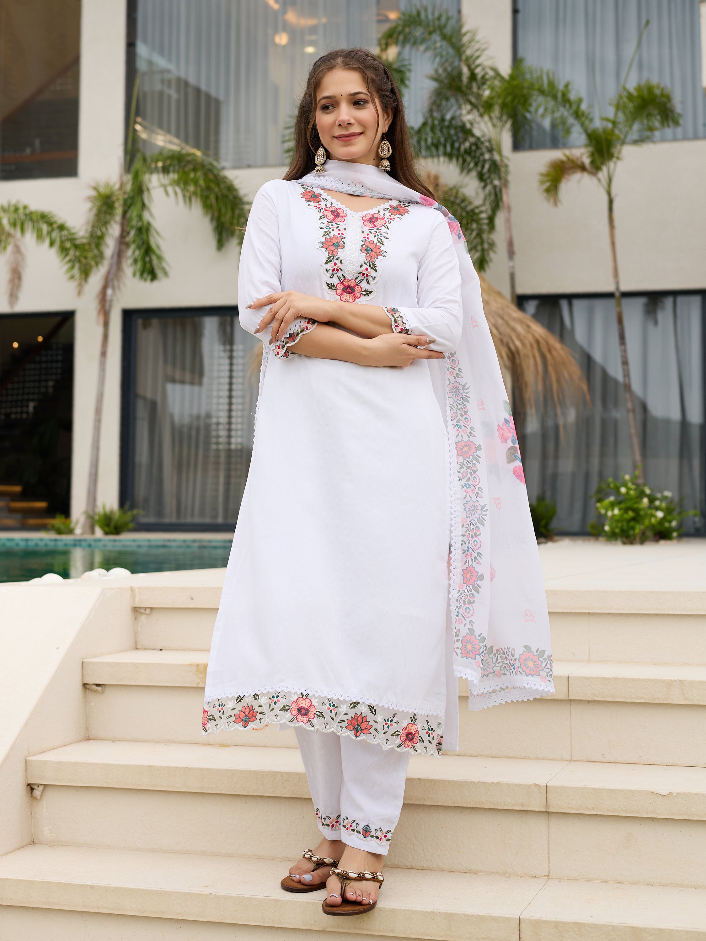 TRENDBUY model elegantly standing on white steps in the white embroidered kurti and pant set with matching dupatta, outdoors.