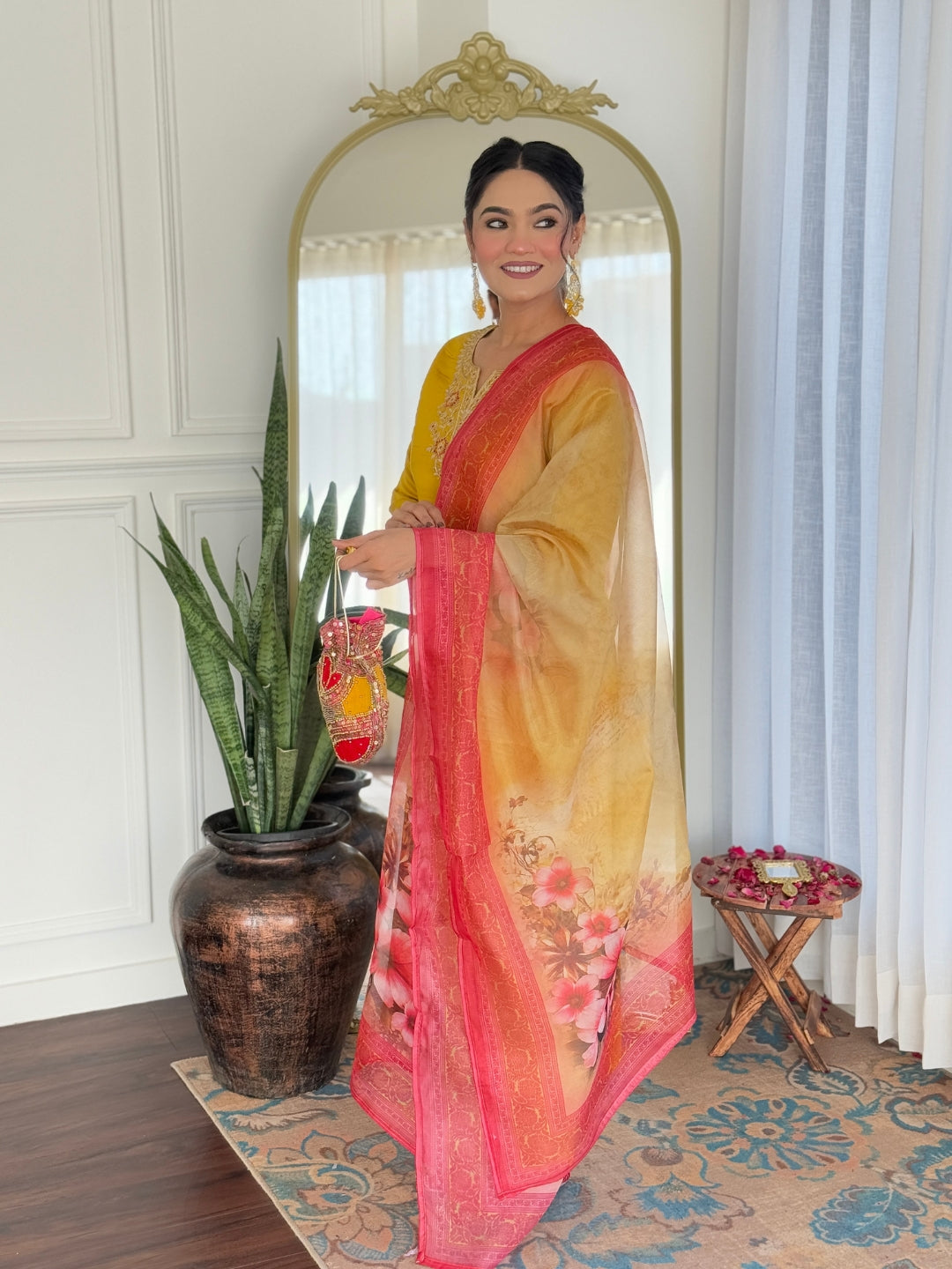 Smiling woman wearing mustard kurti and pant, holding a traditional potli bag, showcasing the floral dupatta indoors