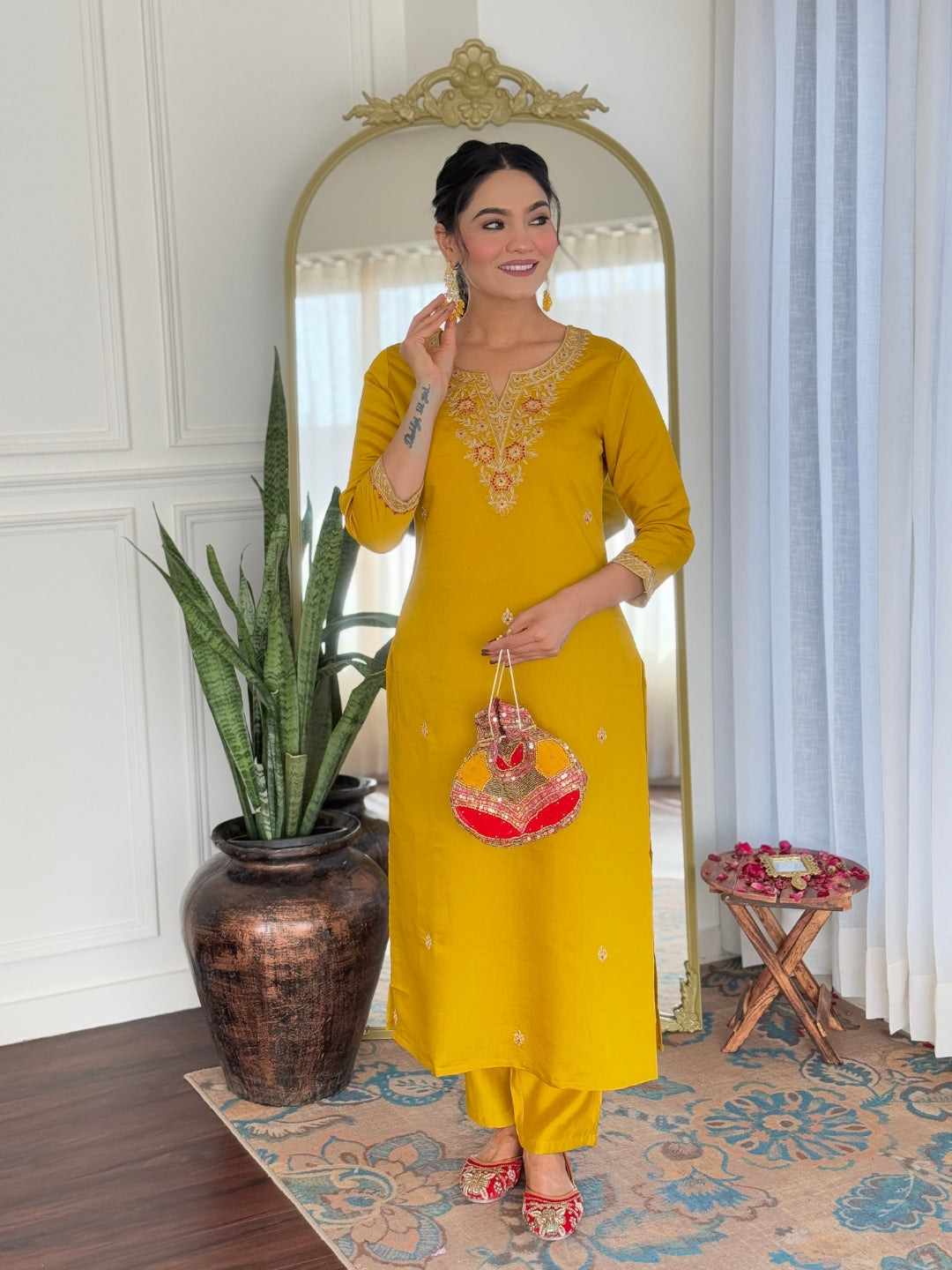 Graceful model in mustard embroidered kurti and pant set, holding a red potli bag, smiling indoors