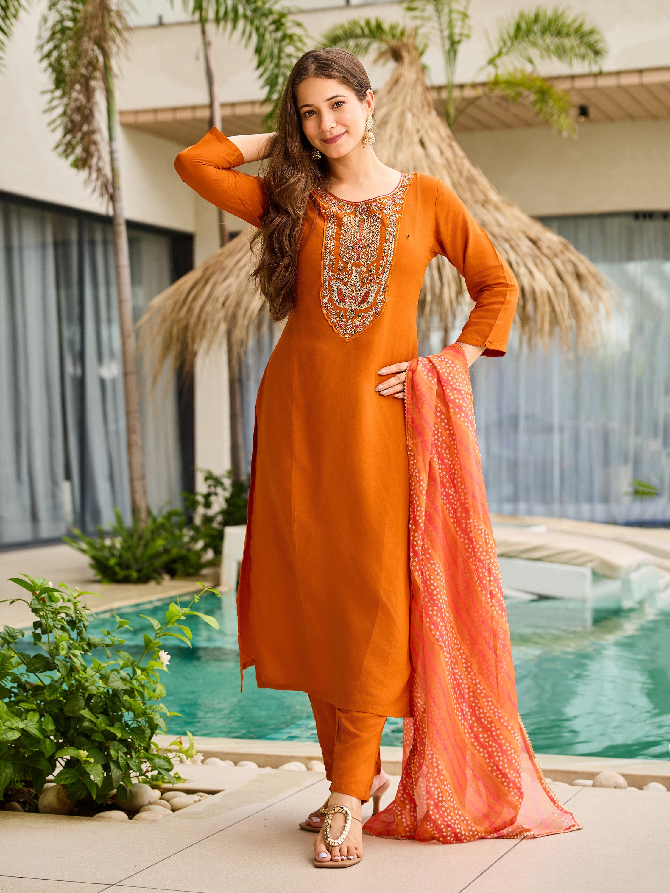 Stylish pose of model in rust orange rayon kurta set, holding the flowing dupatta by the hip next to a pool