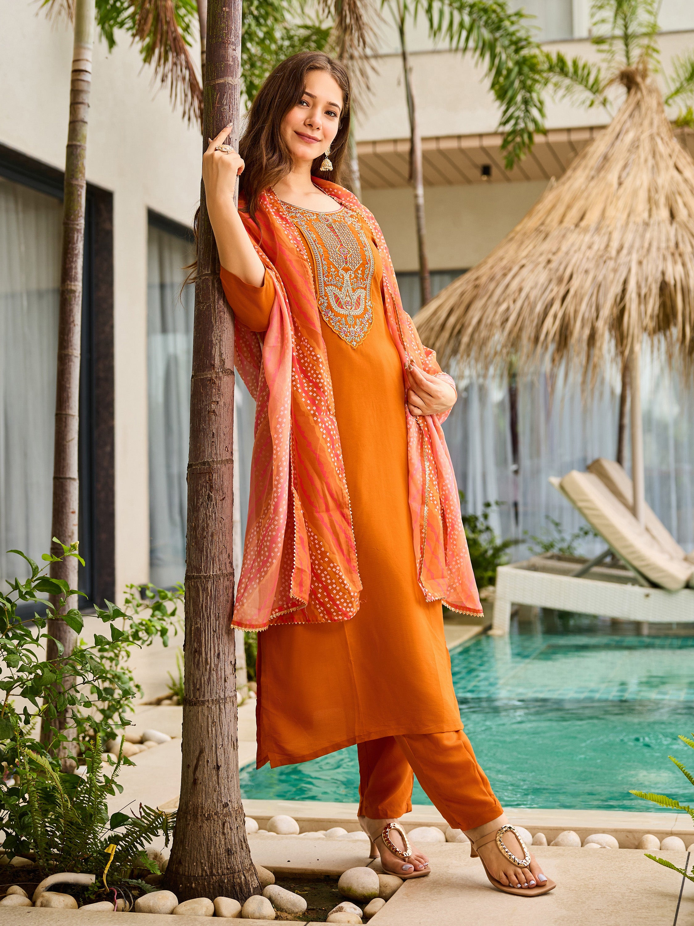 Model leaning against a palm tree in the rust orange embroidered kurta set with the dupatta draped over her arm