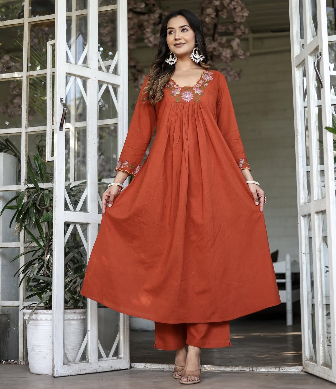 Model in full length, wearing orange rayon slub kurti set, hands holding the flare of the kurti, looking confident.