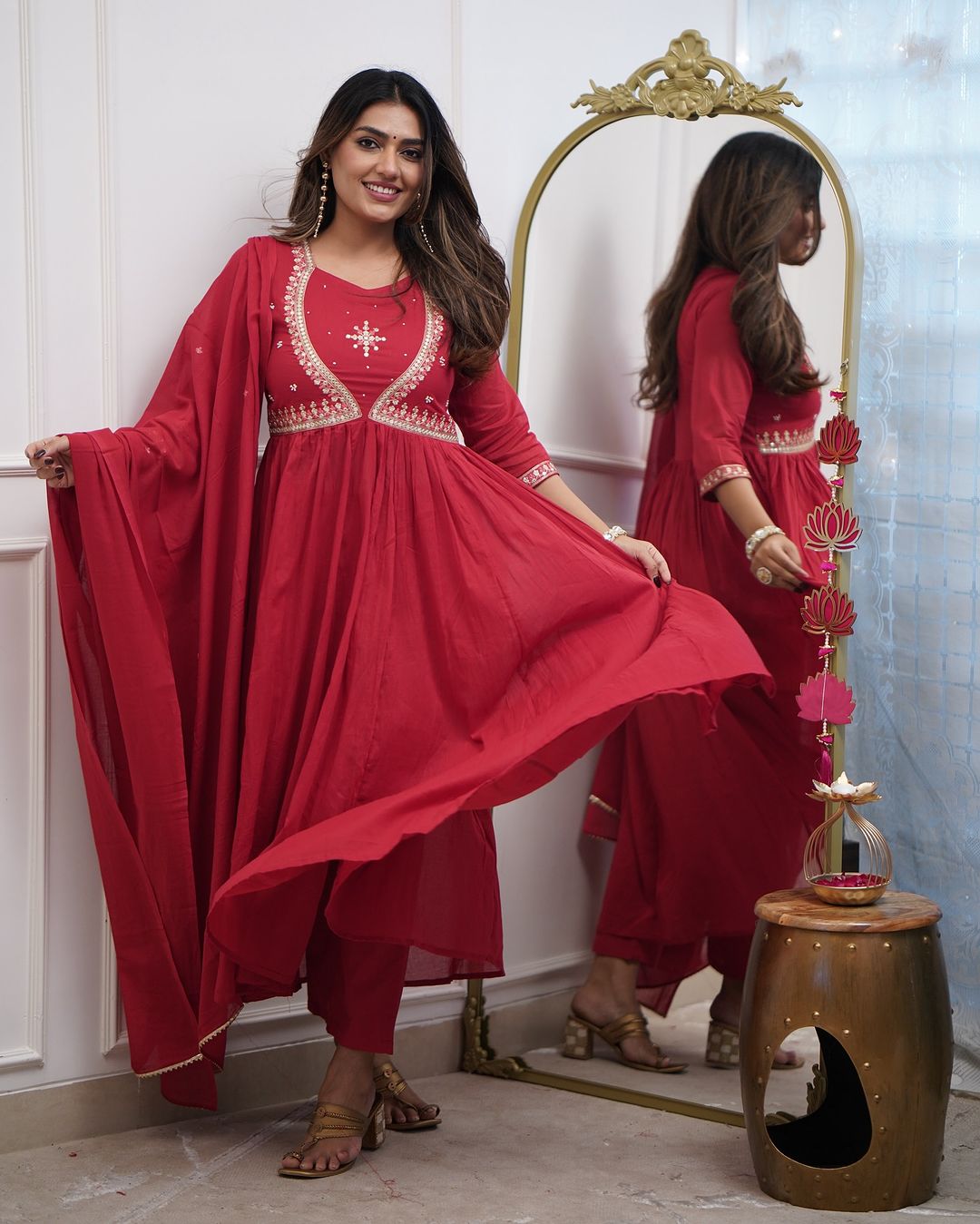 Full shot of a woman in a red Muslin Cotton kurta set, gracefully lifting the kurta to show its flare, posing beside a decorative mirror.