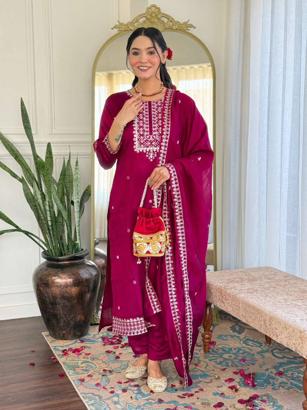 Full-length shot of a woman smiling, posing in a magenta embroidered CP Vichitra kurta set with pant and dupatta, holding a matching potli bag, in a well-lit room.