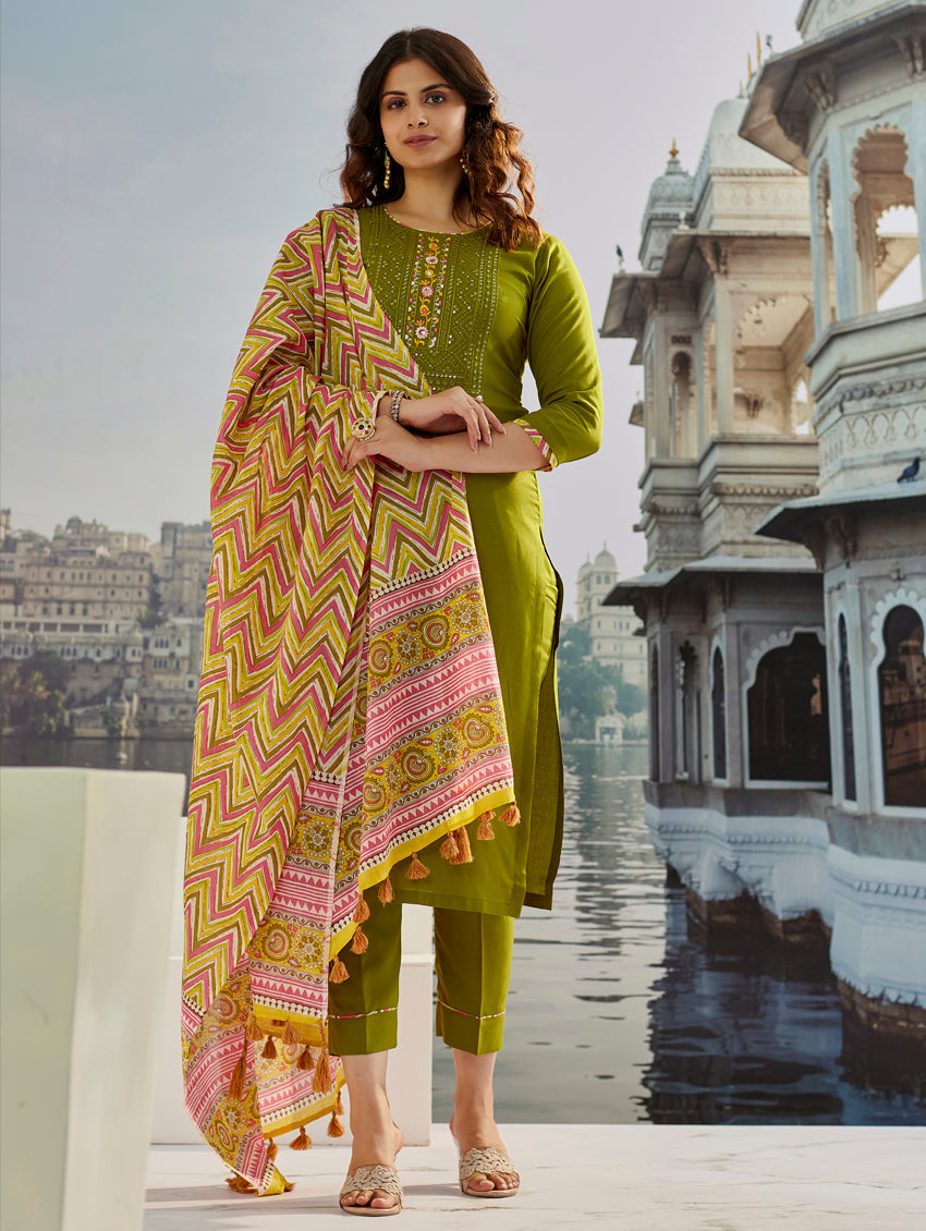 Full-length shot of a model elegantly posing in the TRENDBUY ST-SOMYA olive green Kurta Set, featuring embroidered kurti, matching pants, and a vibrant Chanderi dupatta, set against an architectural background.