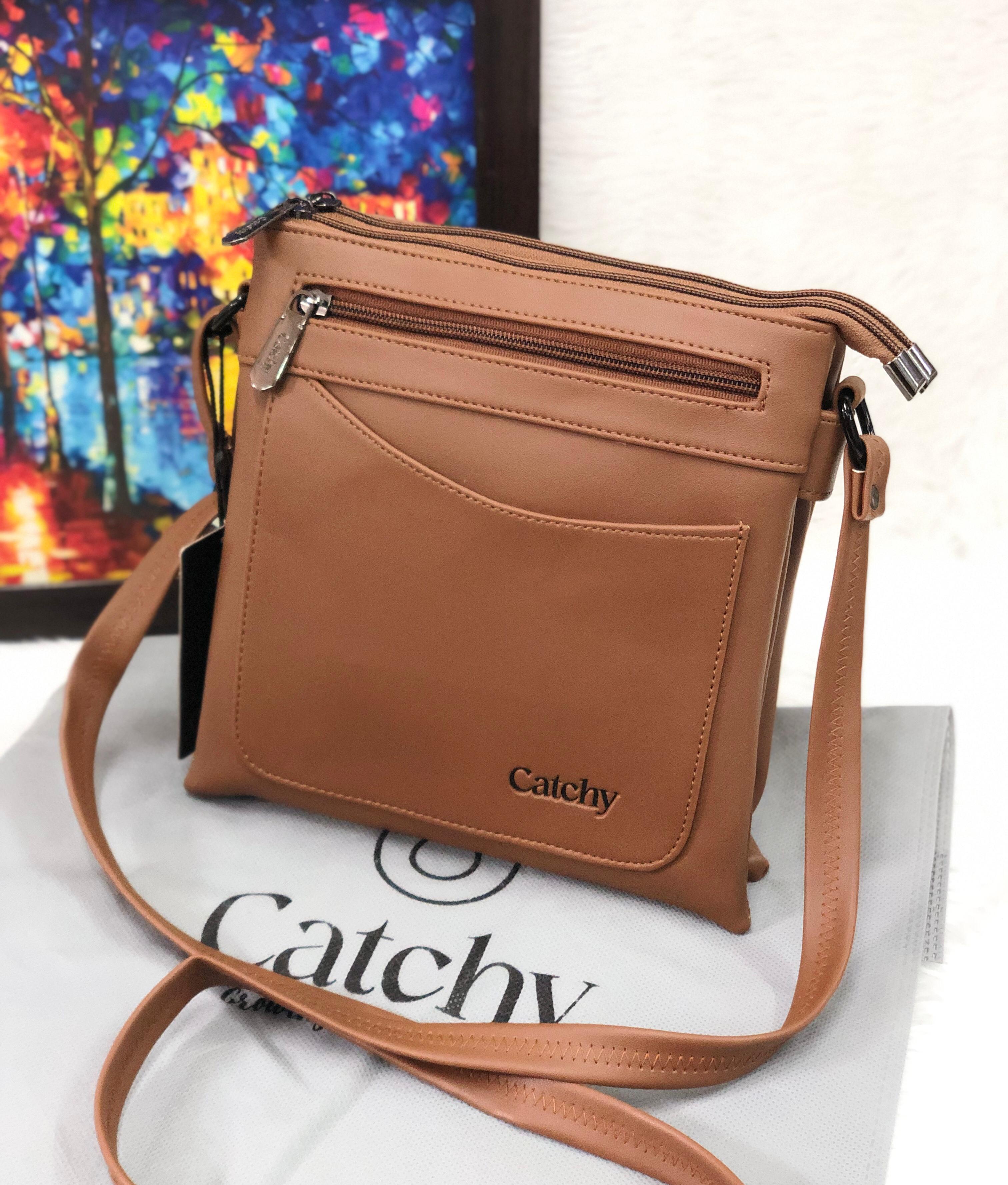 Tan Catchy multi-compartment crossbody sling bag on a white fuzzy surface, with a colorful abstract painting in the background.