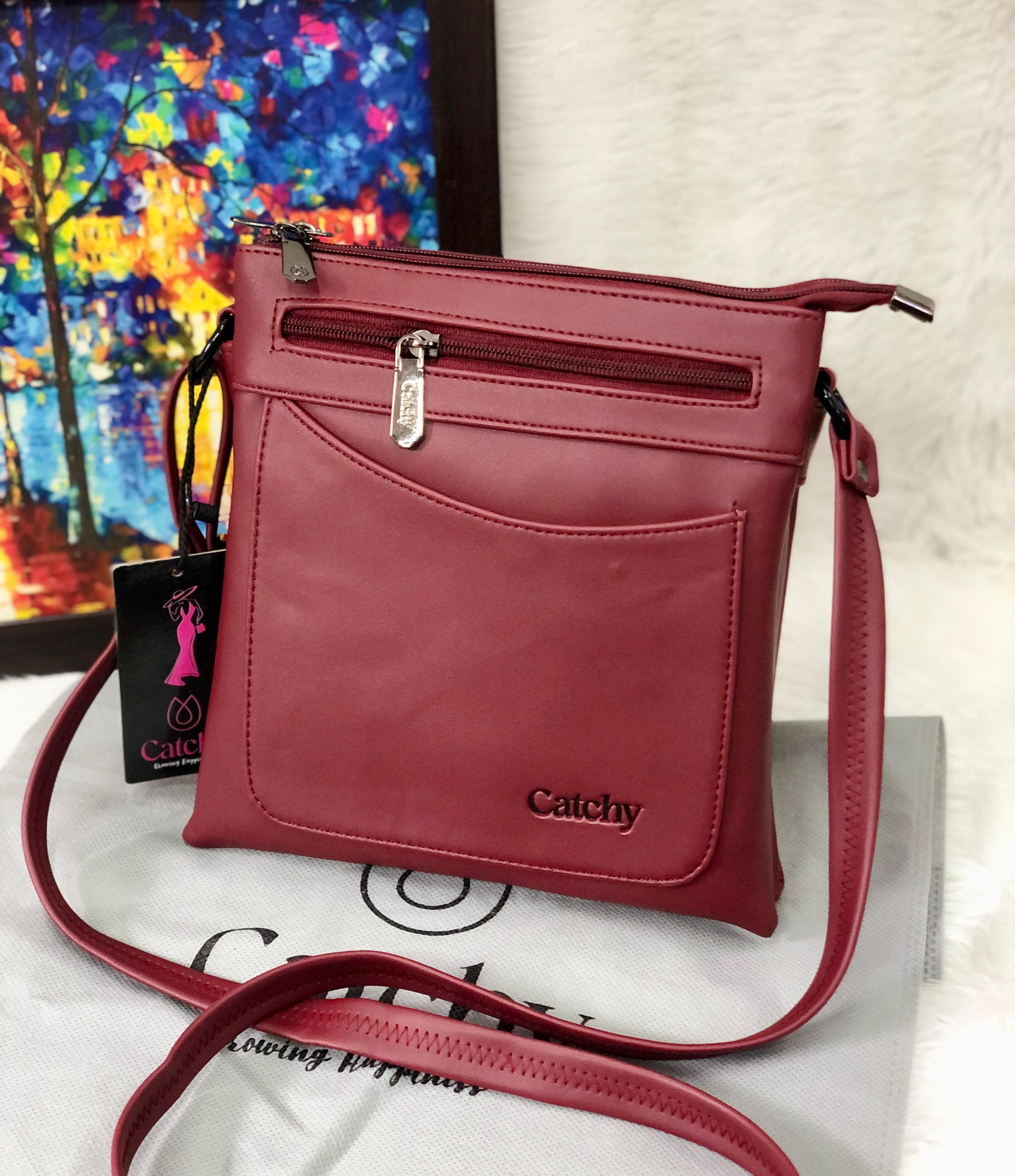 Maroon Catchy multi-compartment crossbody sling bag on a white fuzzy surface, with a colorful abstract painting in the background.