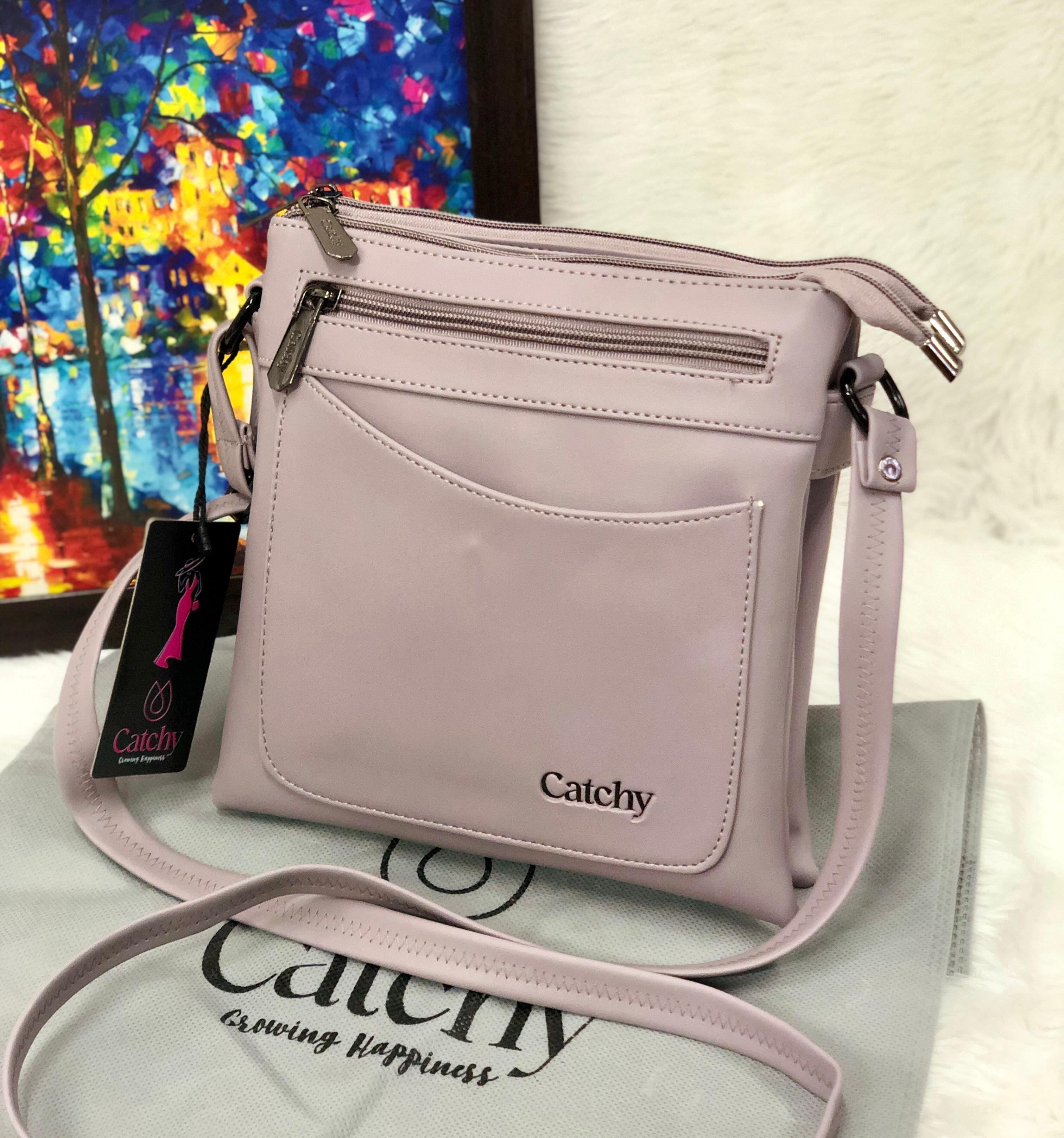 Lavender Catchy multi-compartment crossbody sling bag on a white fuzzy surface, with a colorful abstract painting in the background.